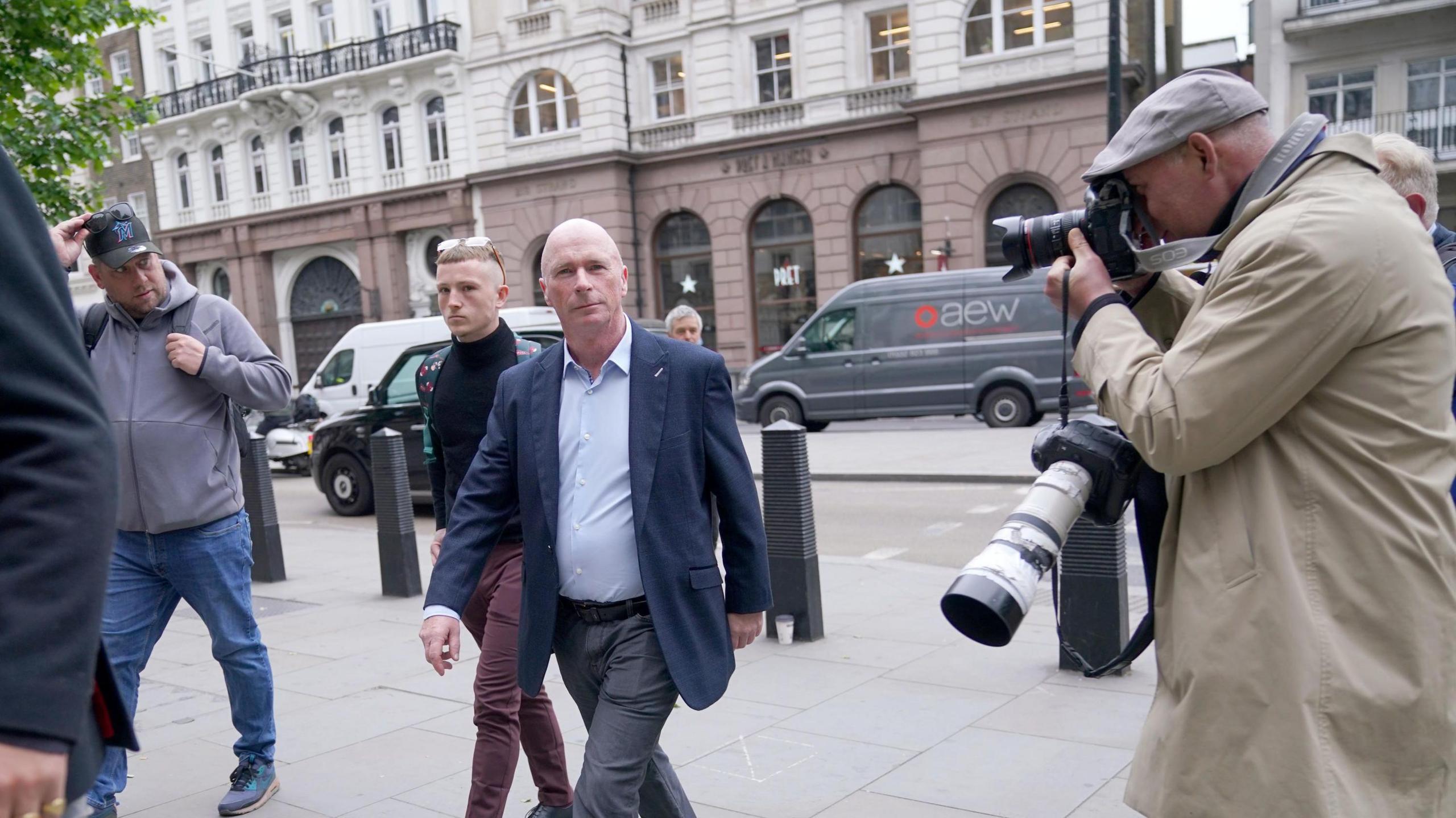 Ray Connolly with very short white hair, wearing a blue jacket and light blue shirt, with grey trousers, walking across a pavement pursued by press photographers. There is a road behind him.