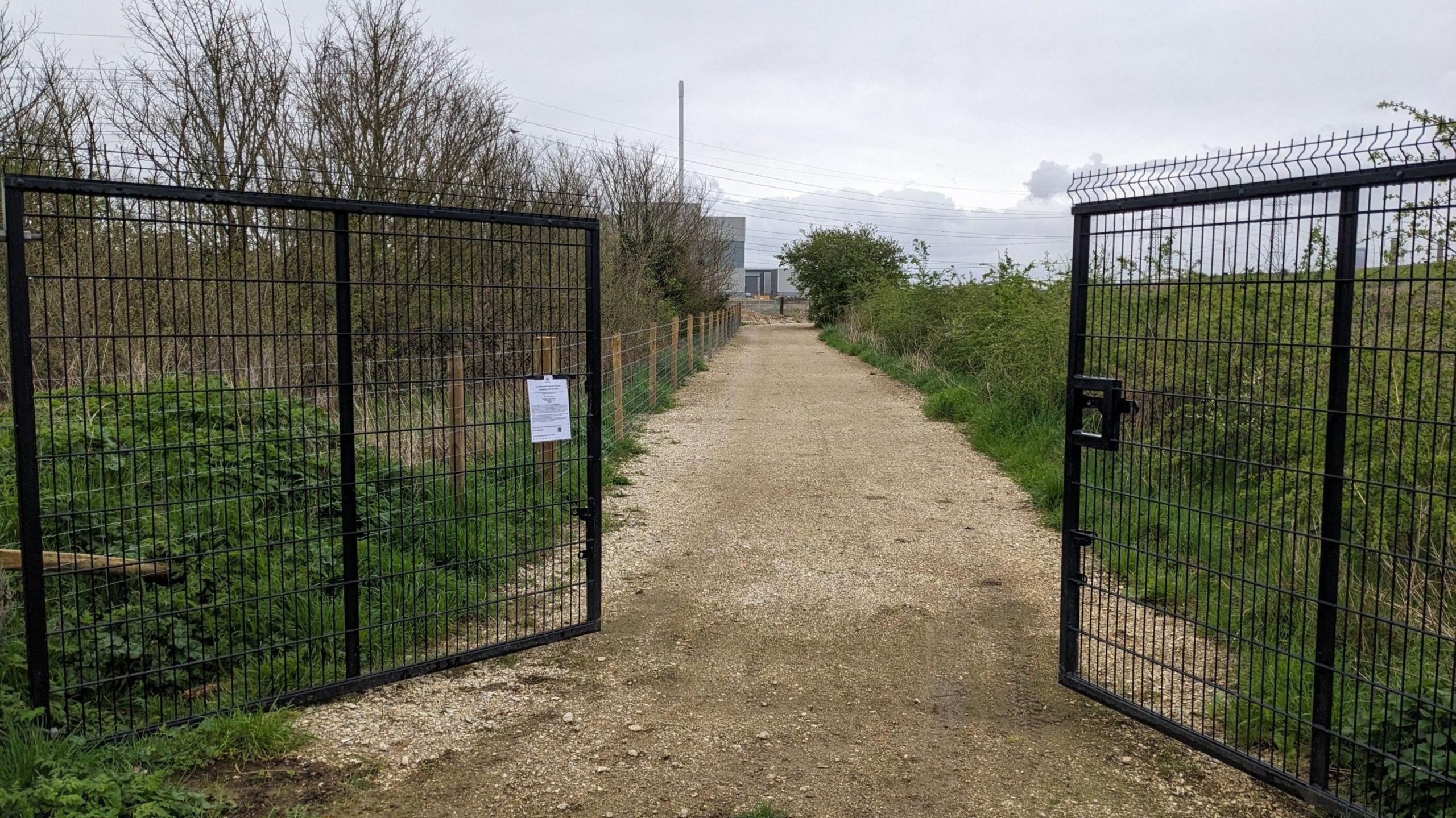 Two black gates are open with a large gravel path down the midland and greenery on either side. 