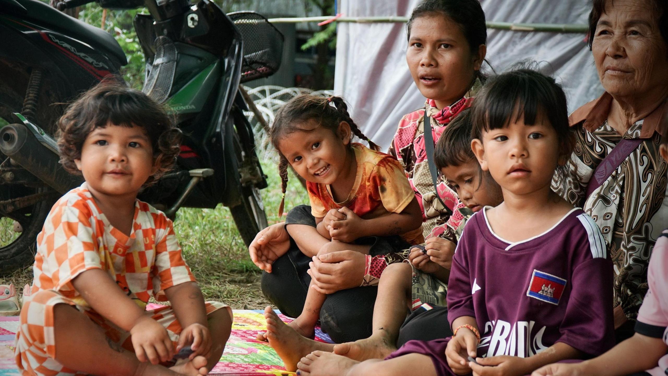 Two women are sitting on the ground, with four children facing the camera, some of them smiling. Two of the little girls are on the women's laps. The other two are sitting on the floor and looking at the camera.  Behind them is a motorcycle.  