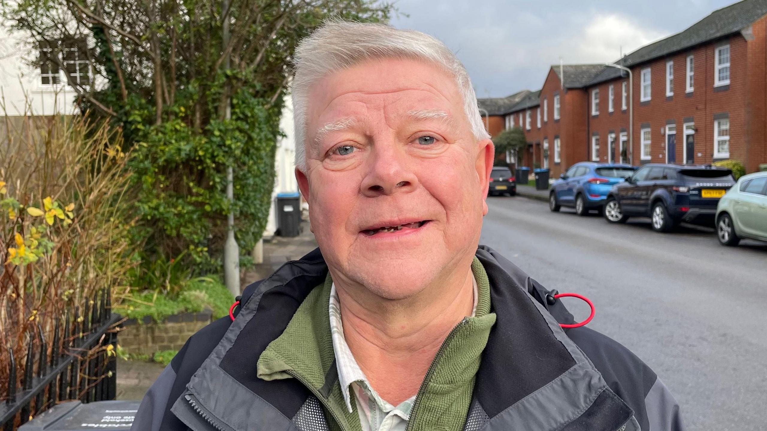 Chris Holroyde looking direct to camera and standing on the side of a residential street. He has grey hair and blue eyes and is wearing a shirt, green half-zip top and a grey and black anorak. To the right of the image are cars parked on the opposite site of the street and a terrace of brick built houses. To his left are some black railings, behind which is a bush.