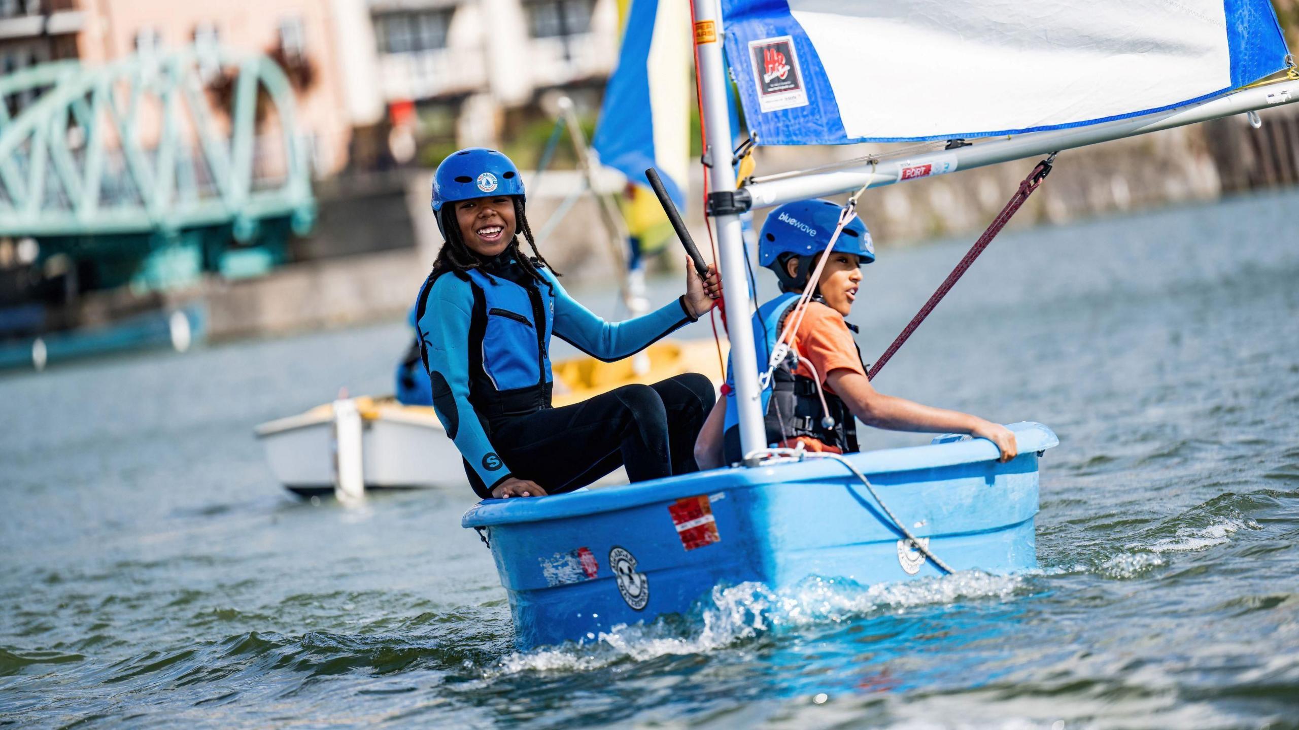 Two young bots on a small blue sailing boat wearing blue clothing and helmets