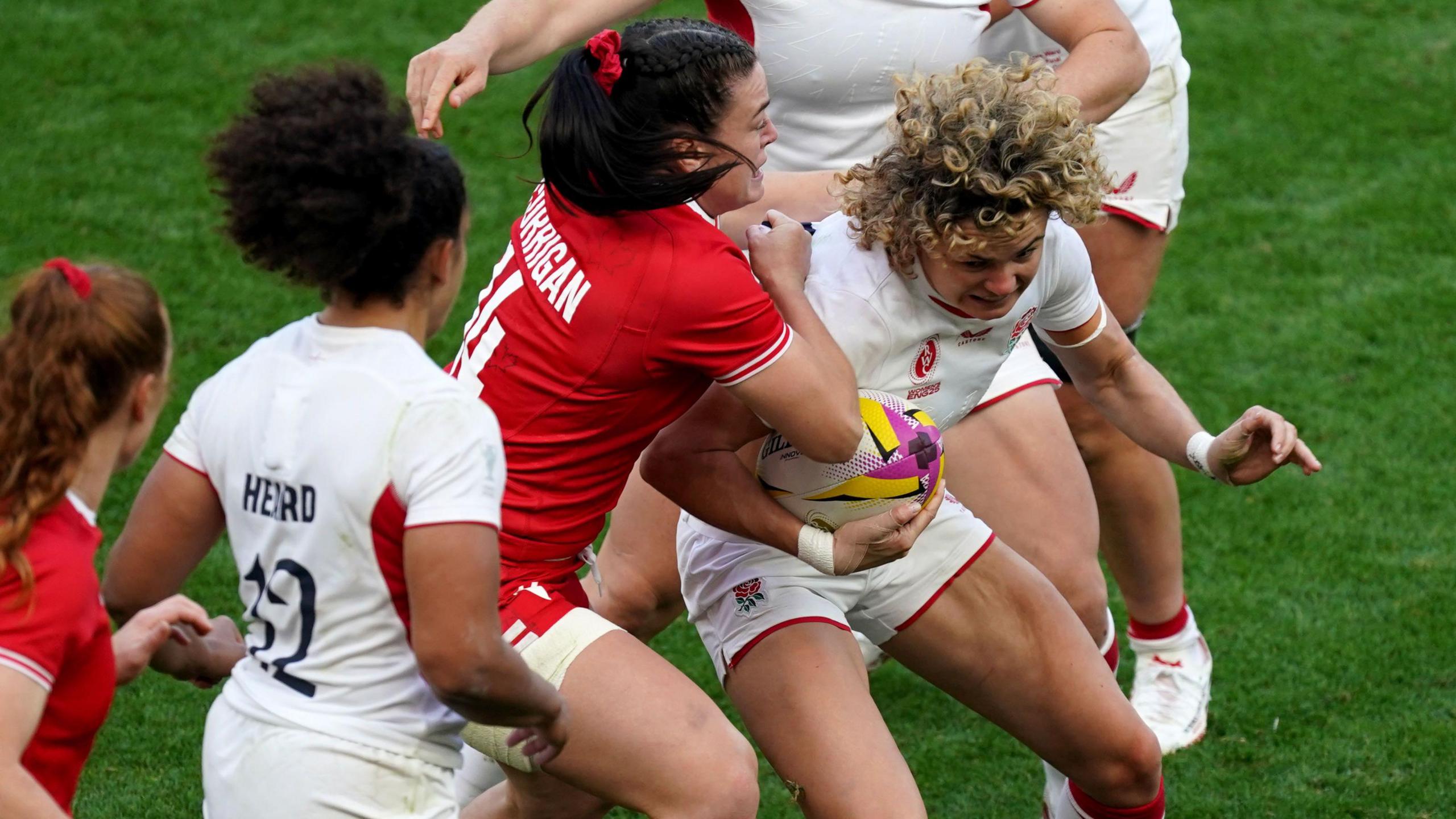 A rugby player wearing a white kit is tackled by another player wearing a red and white kit. Other players are running around them.