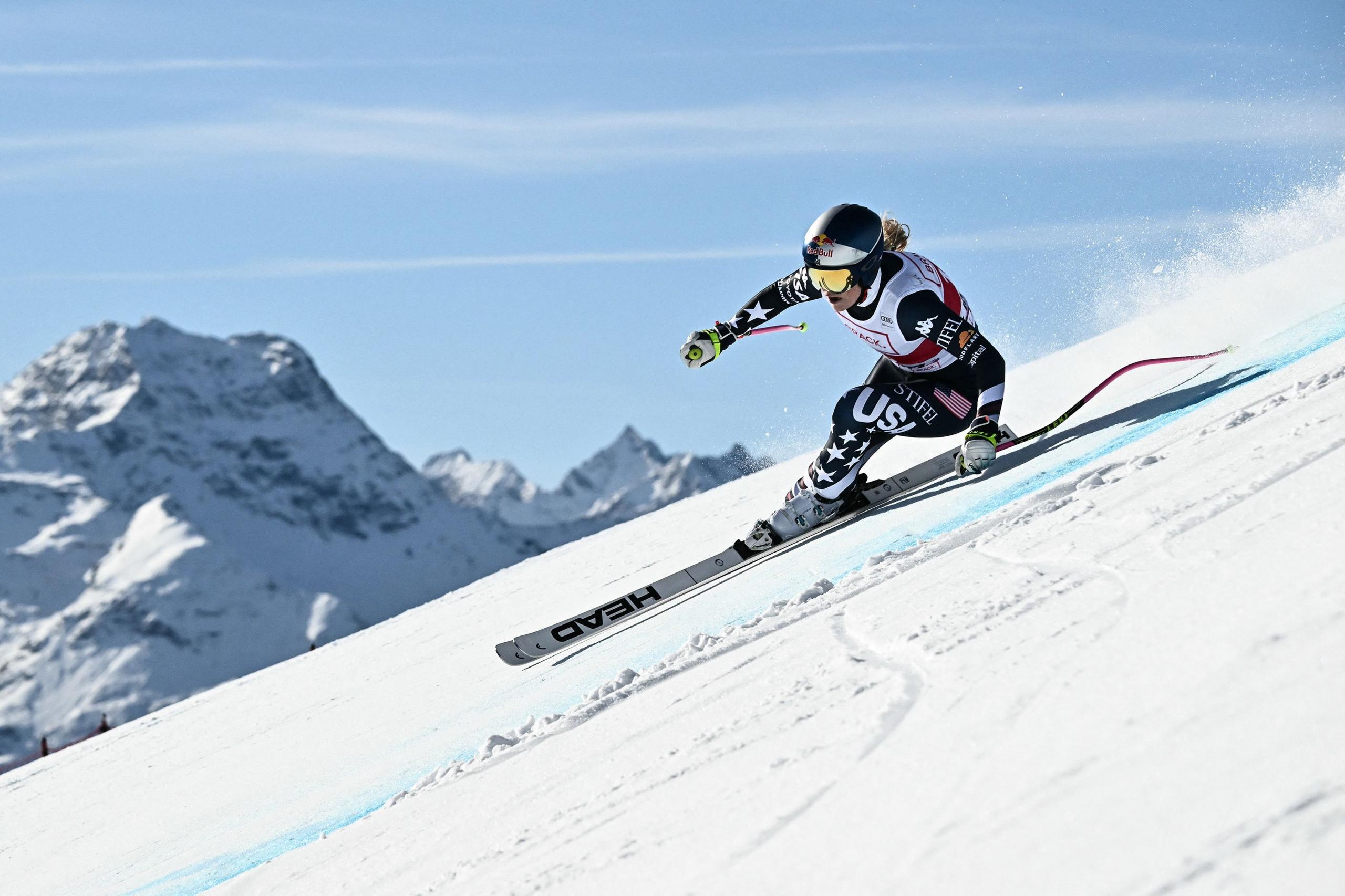 A downhill skier in a racing suit carves sharply through the snow on a steep slope, leaning into the turn with mountains in the background under a clear blue sky.