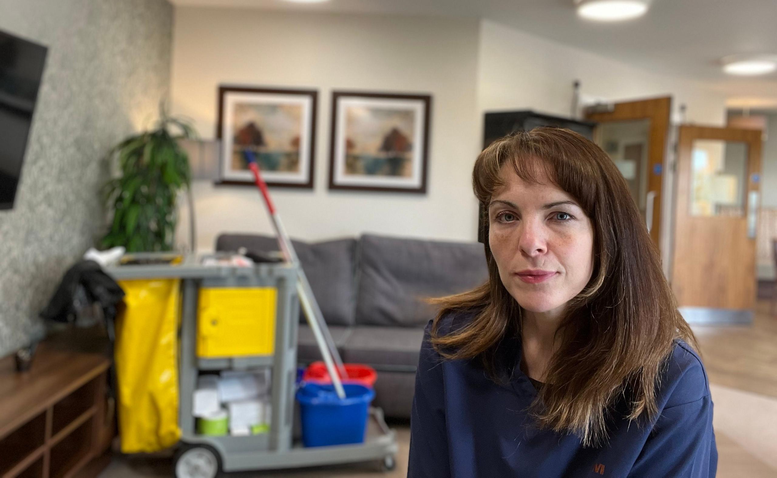 Photo of Catriona in the care home. The photo is a selfie which appears to have been taken with the camera balanced n a table. The background shows the interior of the home with her cleaner's trolley behind. Catriona is dressed in a blue overall.