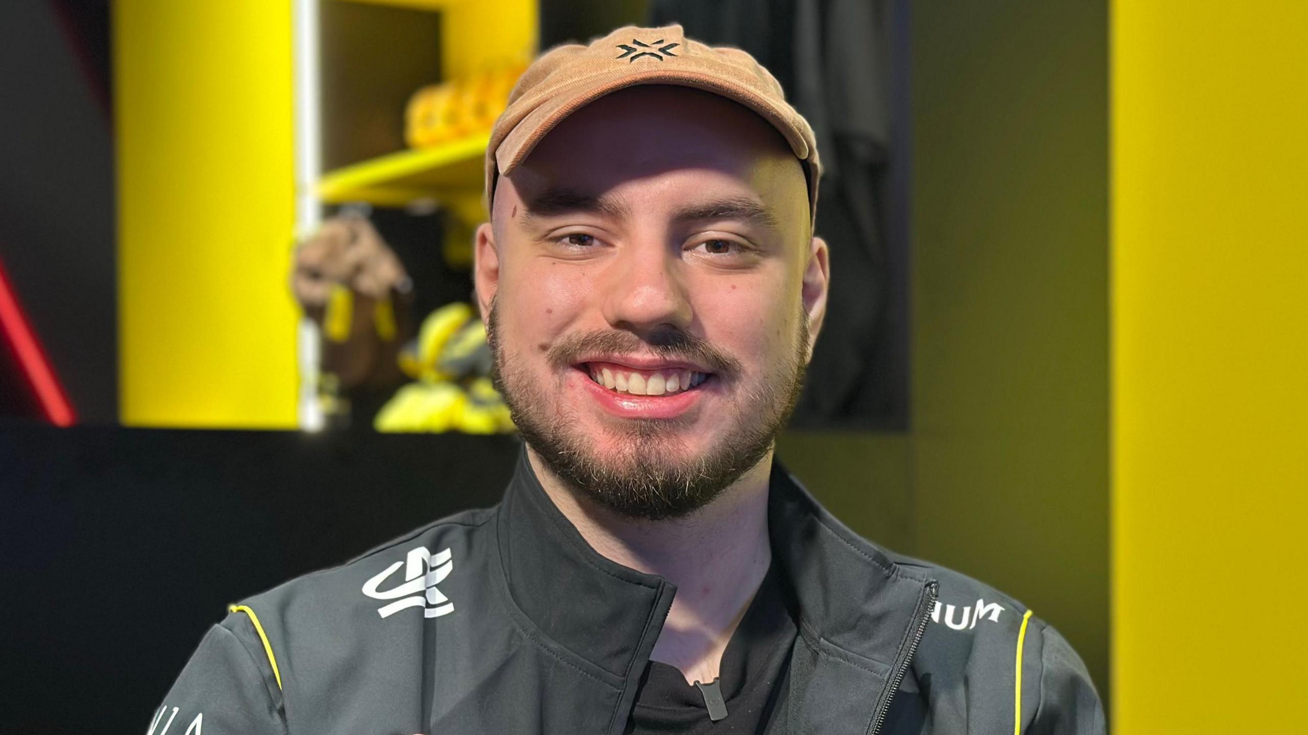 Derke wears a grey zip up fleece with a white logo on it. He wears a brown cap and has a short brown beard