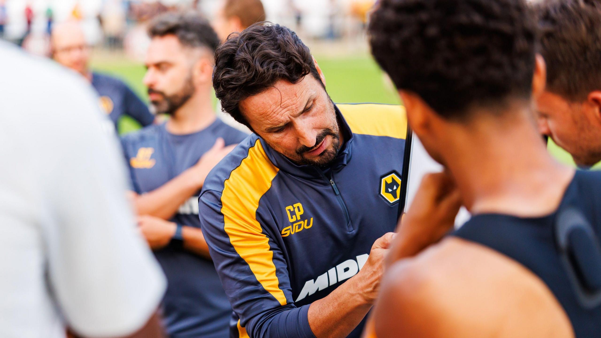 Cesar Peixoto, wearing a black and gold Wolves training top, lays out his tactics to his team using a whiteboard