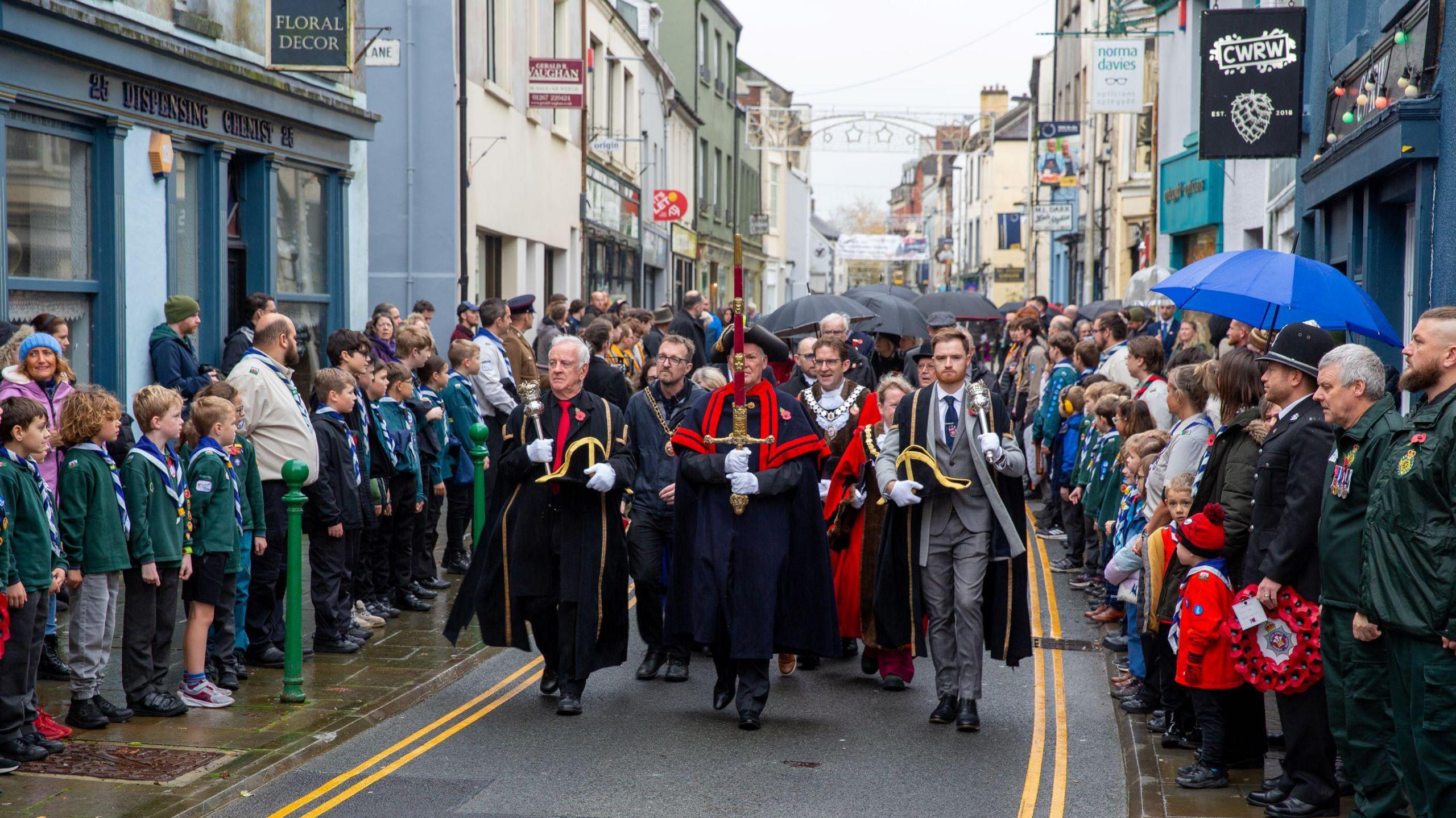 Photograph showing members of the public lining the streets as a parade walks through the road. Some small children dressed in scouts uniforms stand to the left of the road, facing inwards. While soldiers and families stand on the right han side of the road holding umbrellas. A row of terraced shops in different colours line the road and the sky is grey.