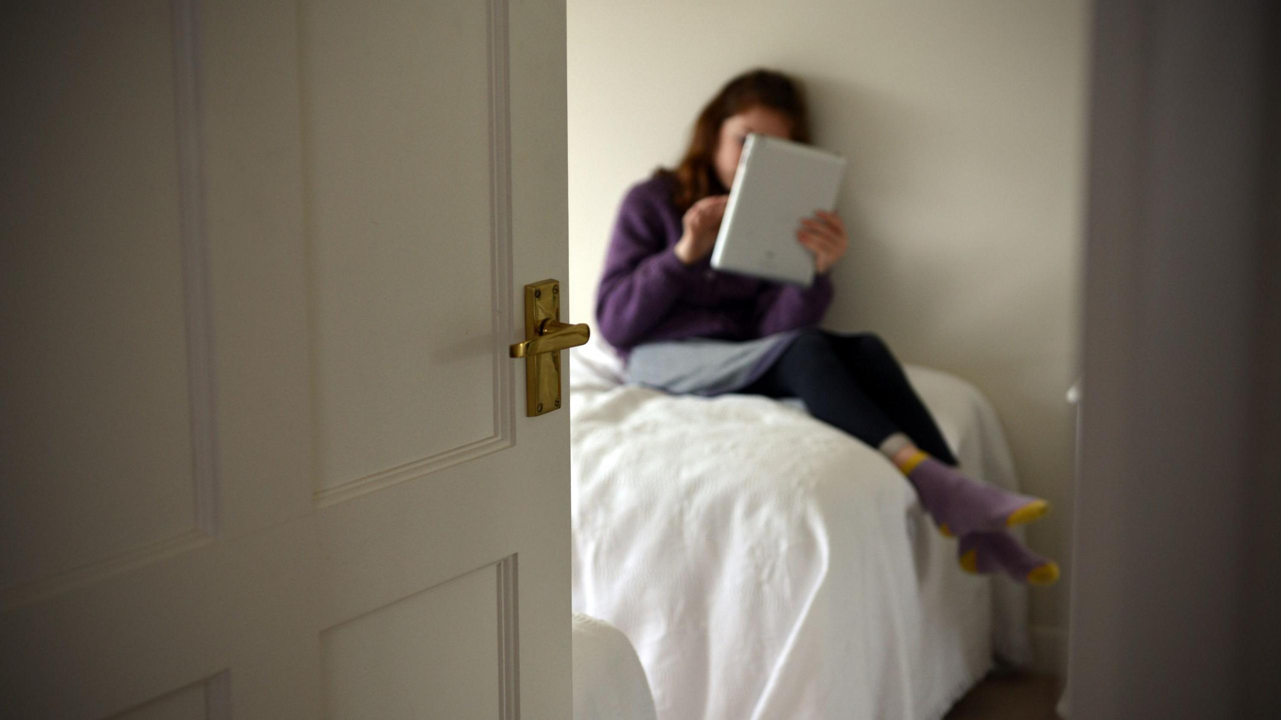 A young girl using a tablet computer on her bed, with the door open.