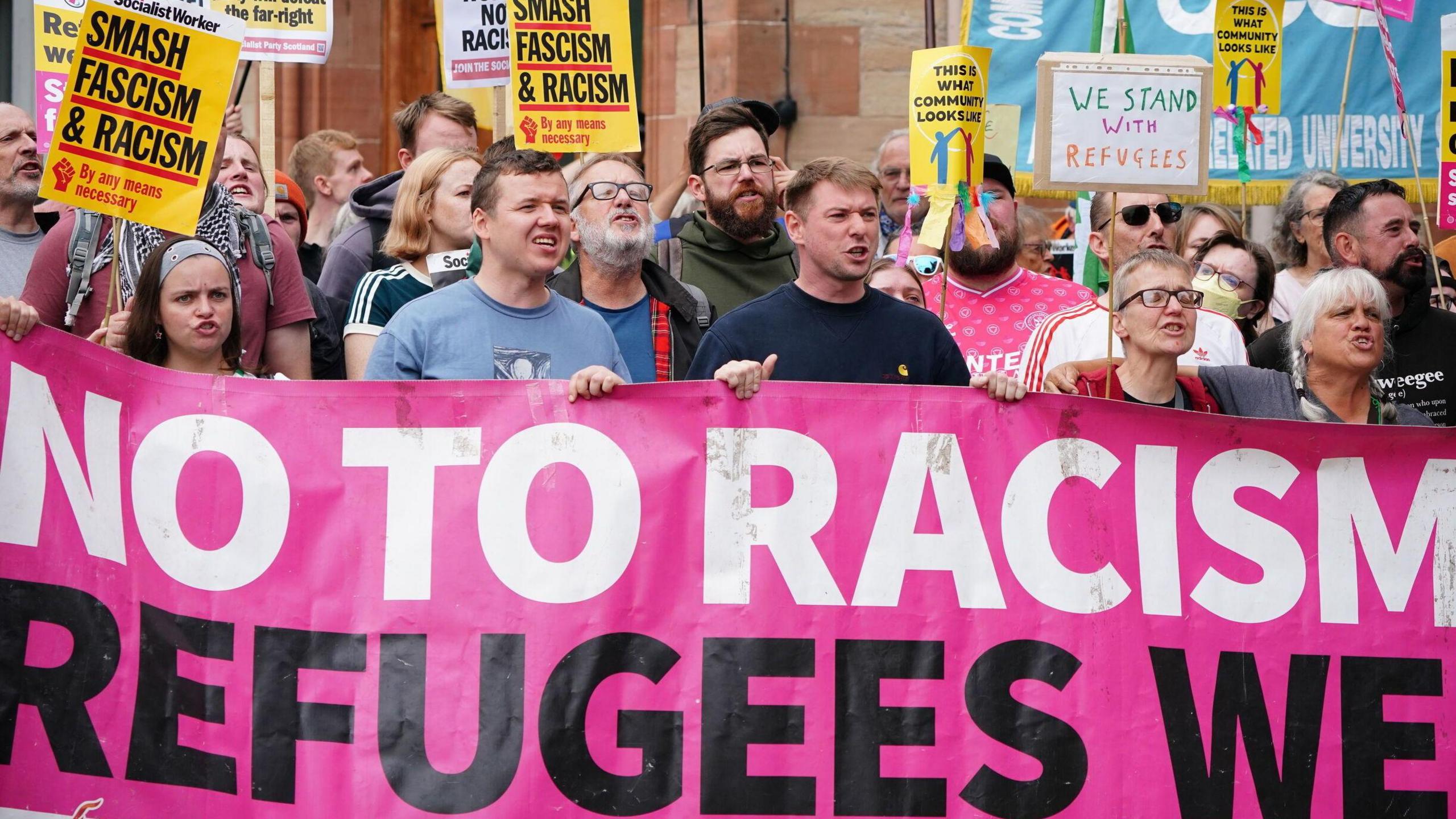 Counter protestors outside the Radisson Blu in Perth. They hold a banner which says "no to racism, refugees welcome" and signs which say things like "we stand with refugees" and "smash racism"
