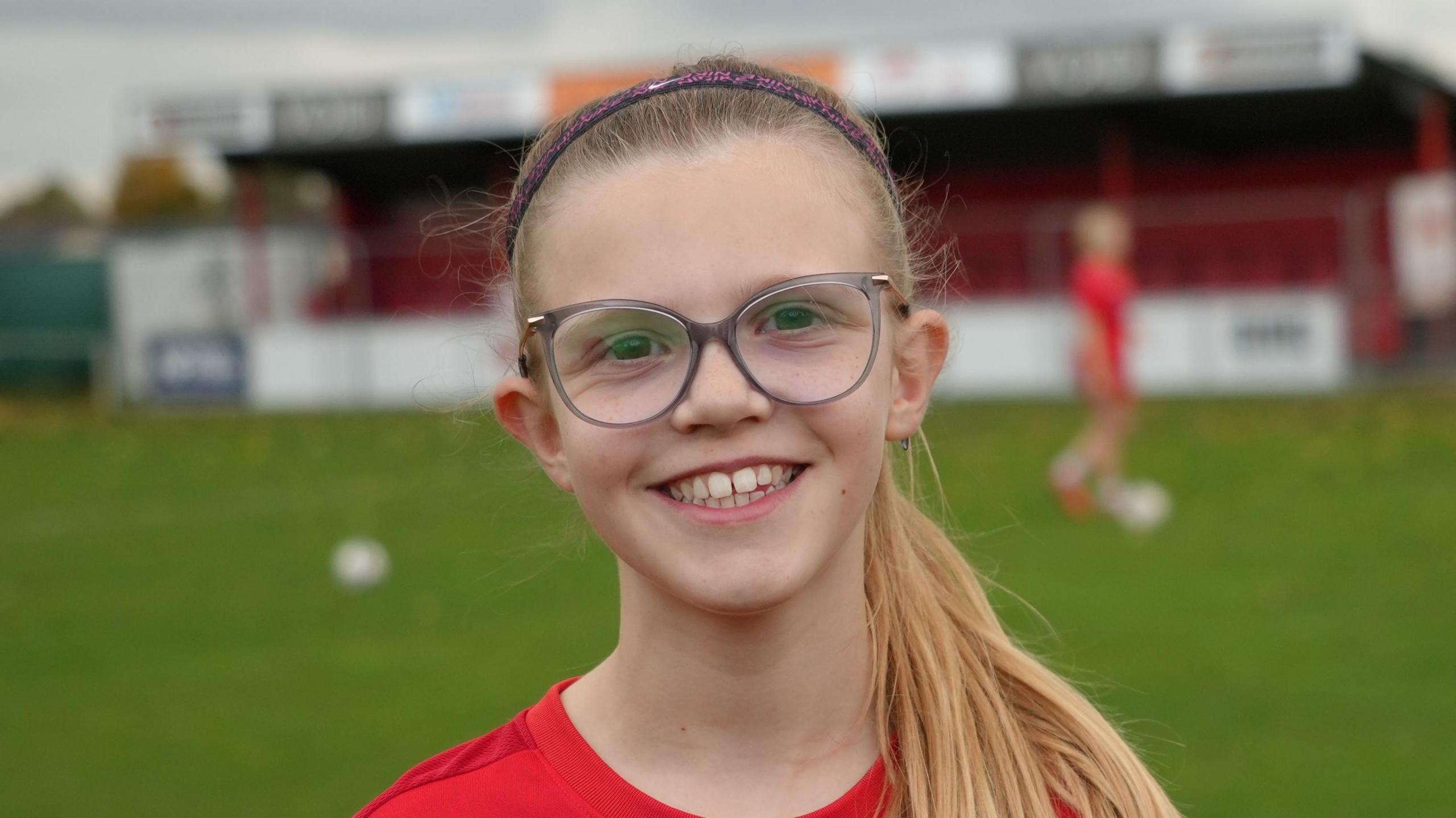 Ruby smiles at the camera while standing on a football pitch in her red football shirt. She has long blonde hair that has been tied up with a purple headband and glasses.