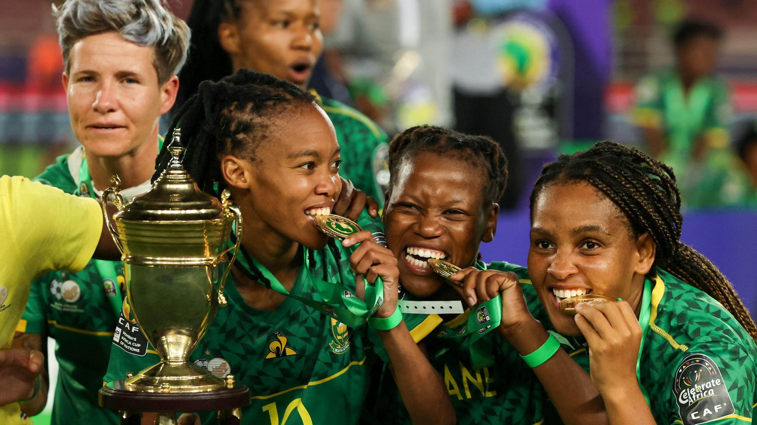 Three South Africa players, wearing green shirts, bite their winners' medals as the pose alongside the Women's Africa Cup of Nations trophy