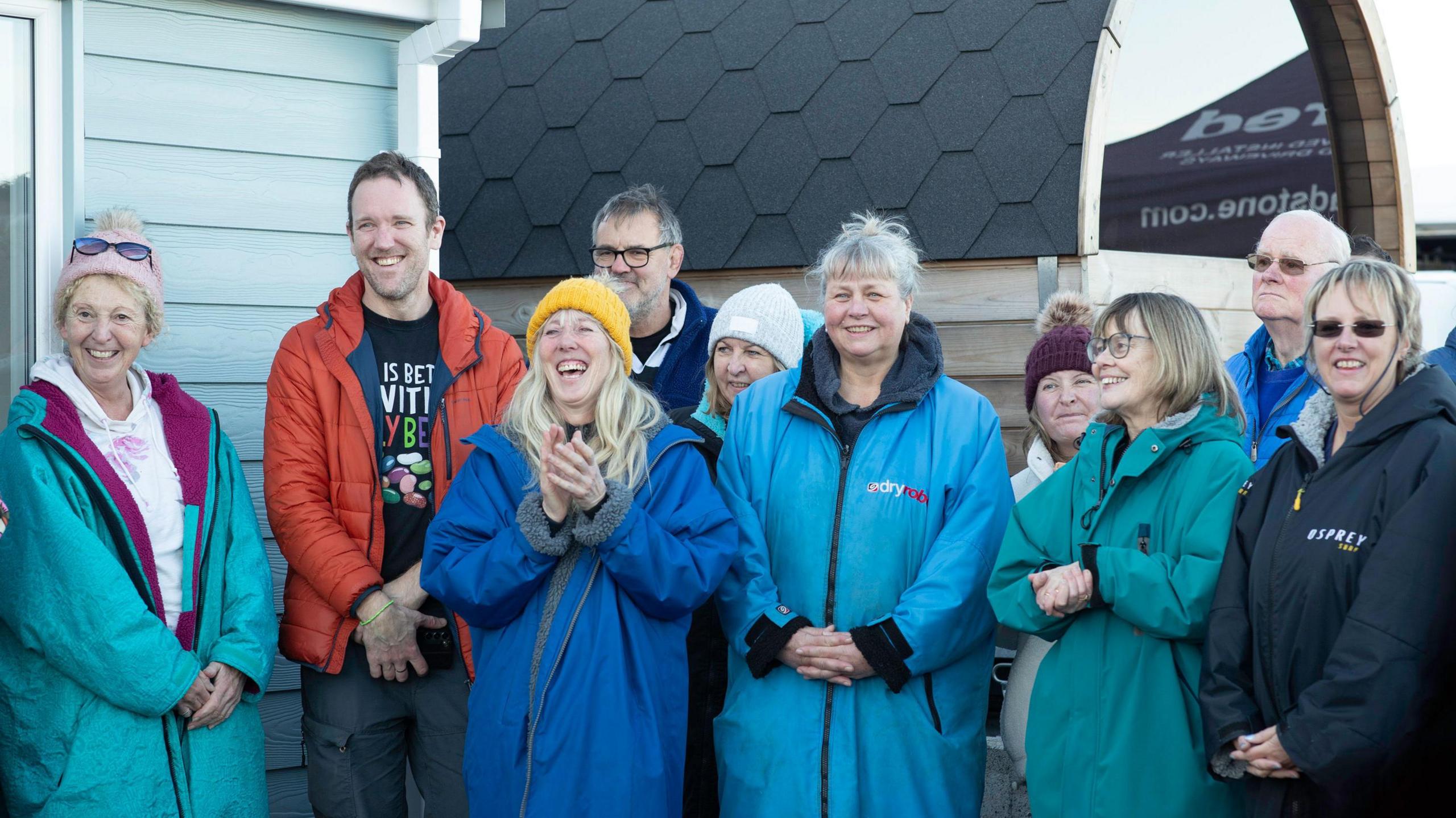 A group of people in multicoloured dry robes, hats and sunglasses gather for the ceremony next to the shelter and nearby beach sauna.