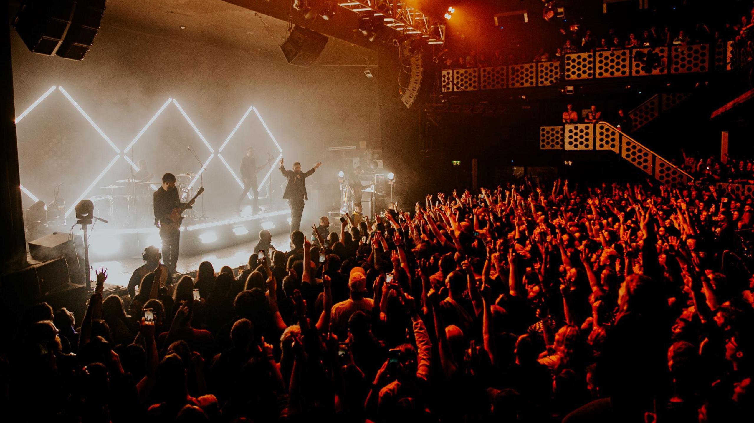 Rock band You Me At Six perform on stage at the O2 Academy in Bristol. The picture is taken from the side of the venue showing the band on stage backlit by white lights and the standing area packed with fans with their hands in the air.