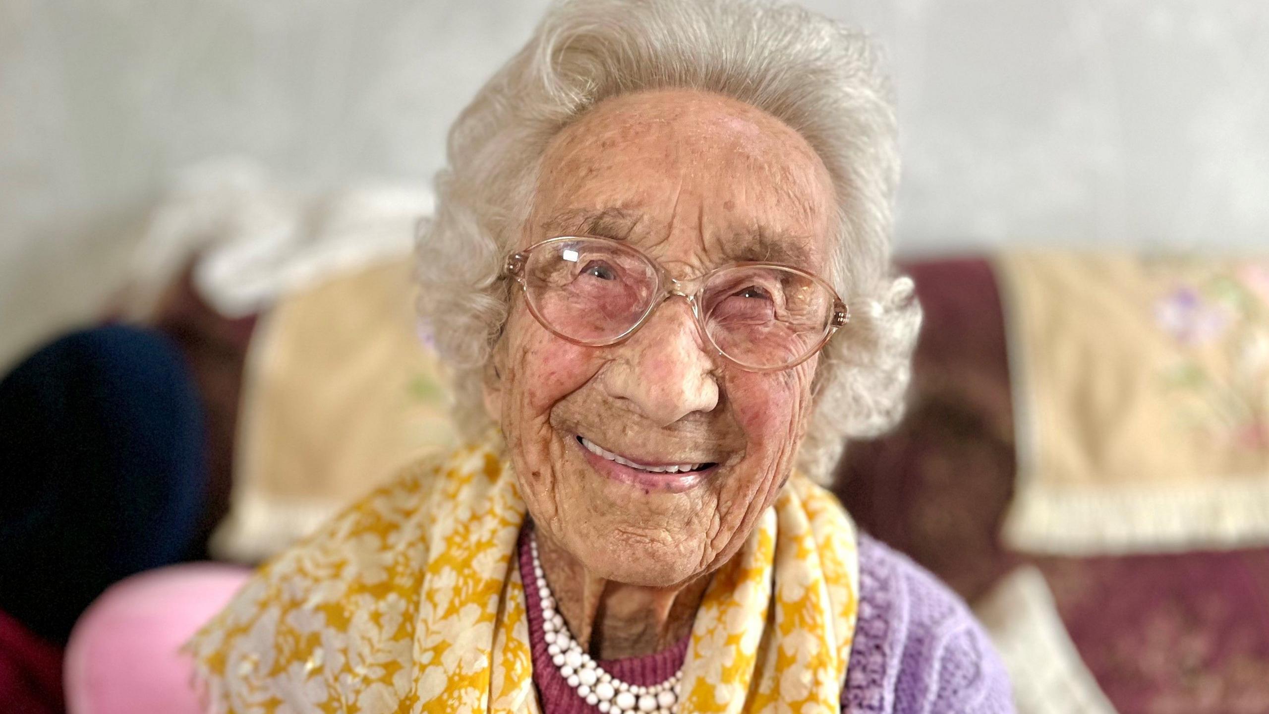 Genna Brown is smiling happily into the camera. She is dressed in brightly-coloured clothes and has glasses and short and curled grey hair. She has a pearl-style necklace on, a yellow scarf, magenta top and purple cardigan.