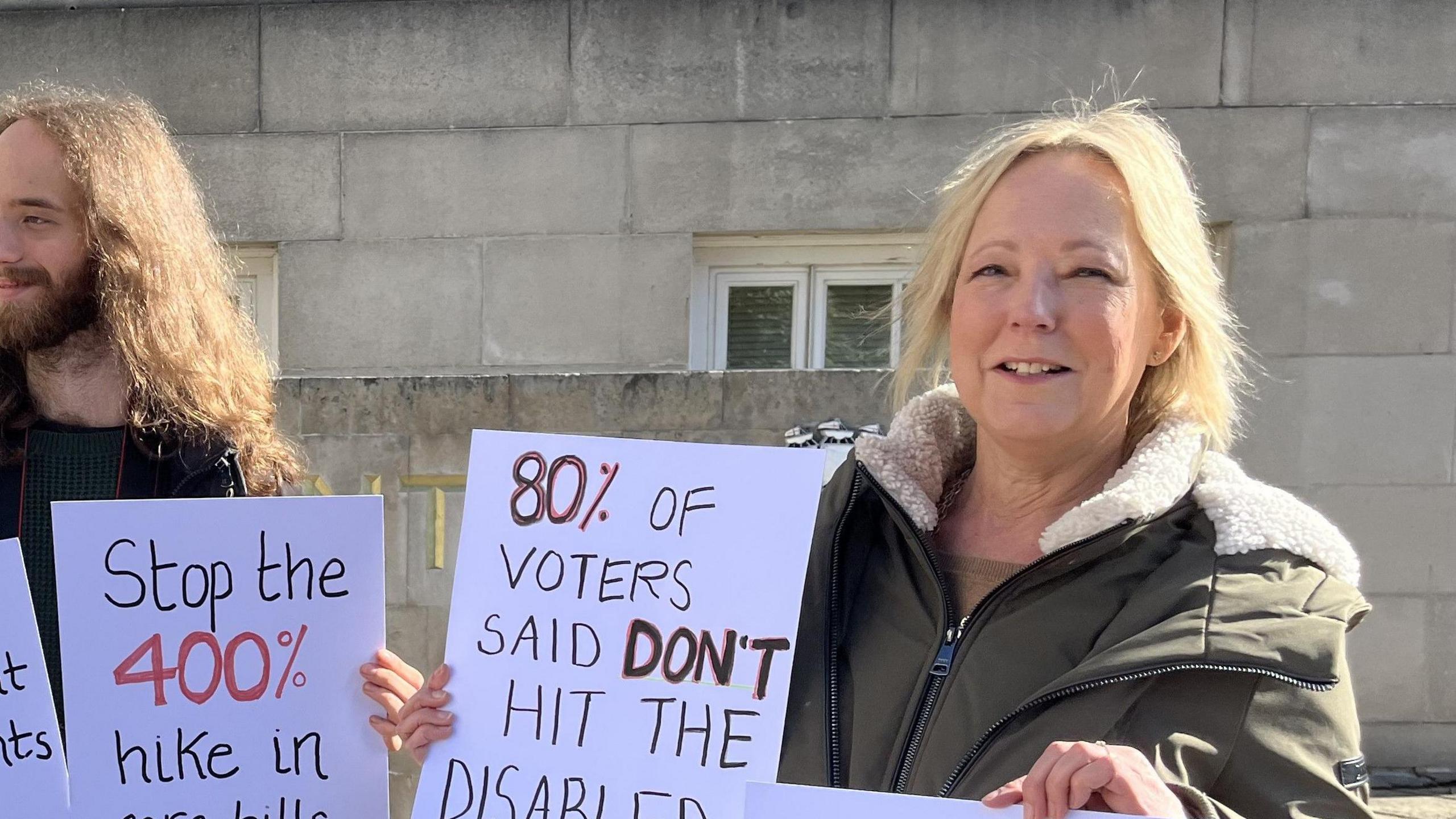 A man with a beard and a blonde lady hold protest signs which read "stop the 400% hike in care bills" and "80& of voters said don't hit the disabled"