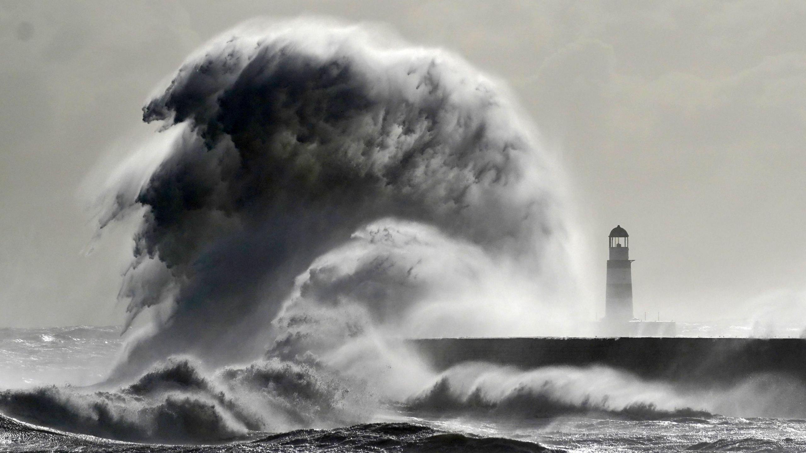 A huge wave rises from the sea, crashing into a harbour. There is a lighthouse to the right which is dwarfed by the wave.