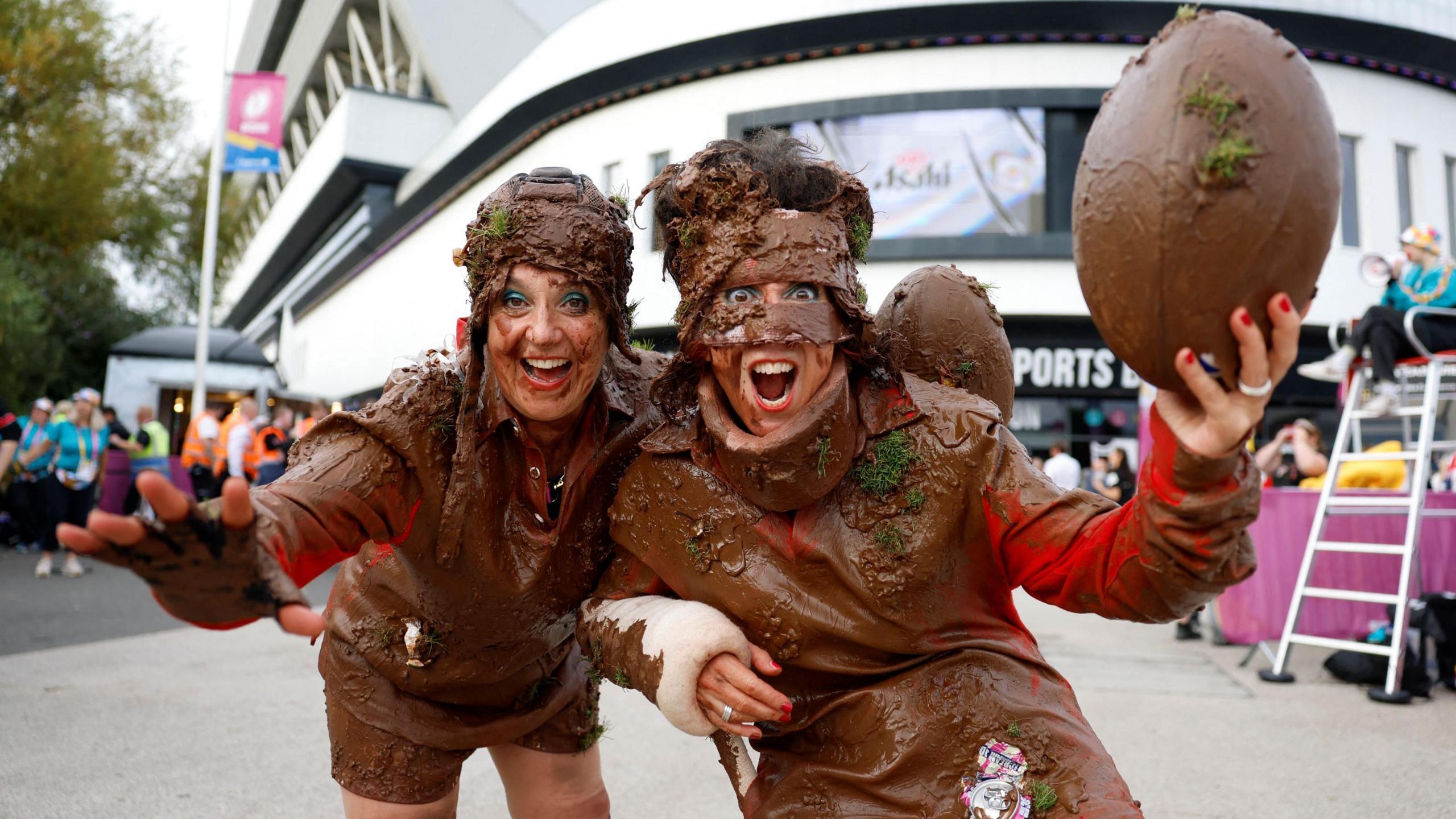 Two female rugby fans smile as they pose for the camera outside Ashton Gate Stadium. They are wearing fancy dress outfits which involve lots of material, which is covered in mud. One of them is also holding a muddy rugby ball in one hand. The picture was taken before the World Cup semi-final match between Canada and New Zealand