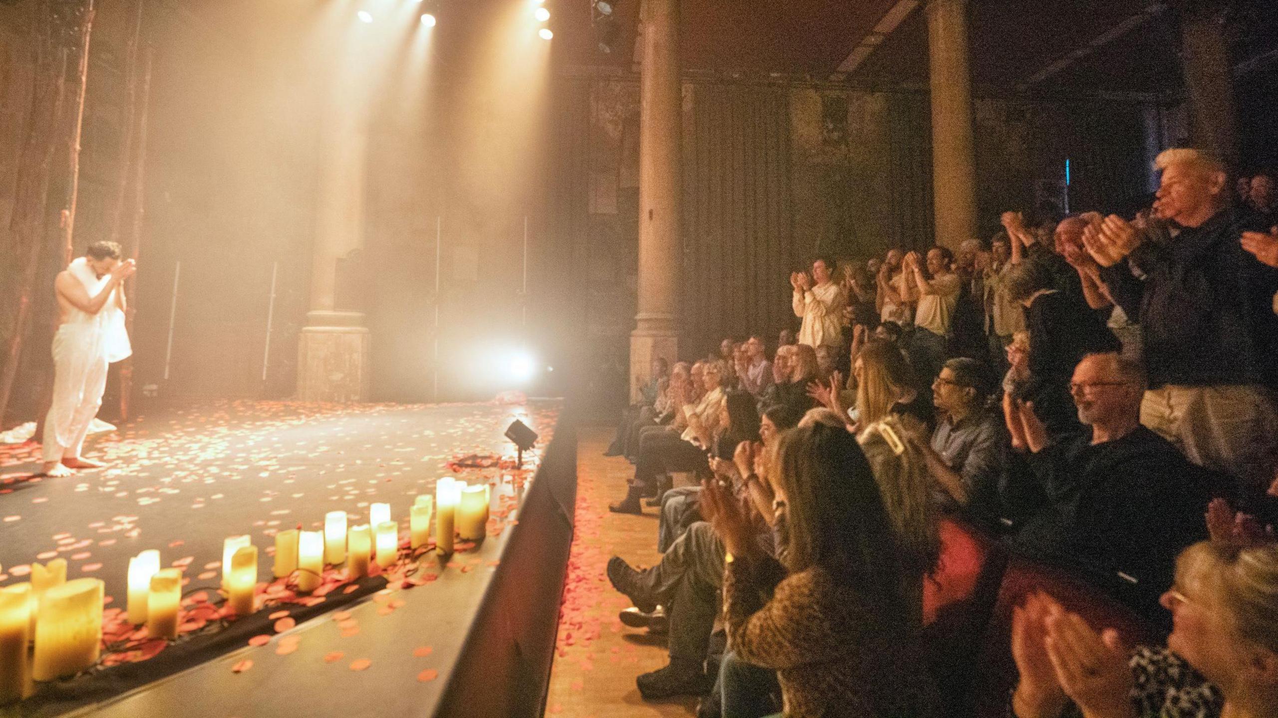 A performer in a white outfit stands on the stage at Bristol Venue The Mount Without as he takes the applause of the audience at the end of a show called Songs of the Bulbul