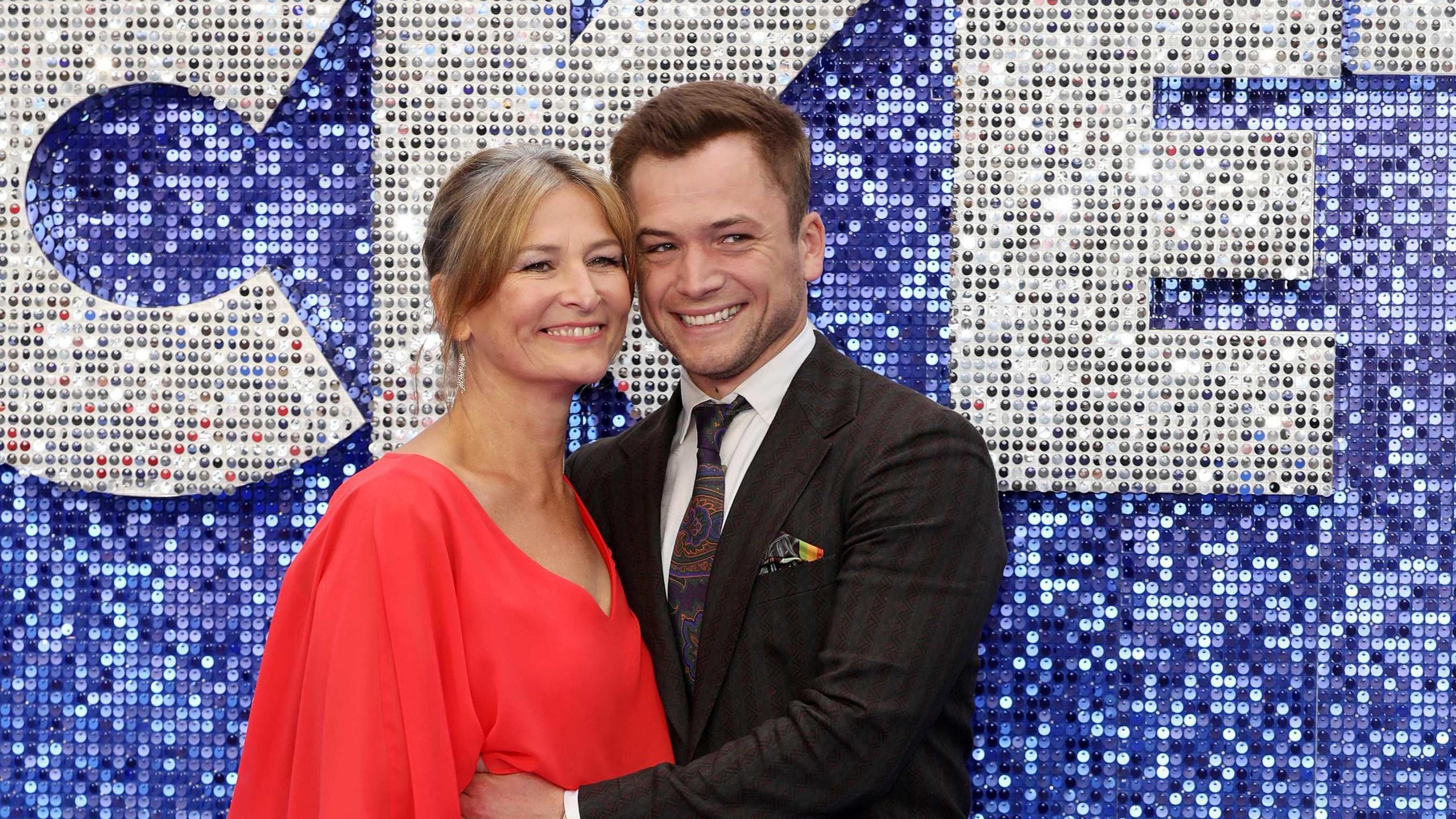 An older blonde woman in a red dress and Egerton with brown hair and wearing a suit. They're on a blue "red carpet" and it has a blue sequin background with silver sequin words behind them.