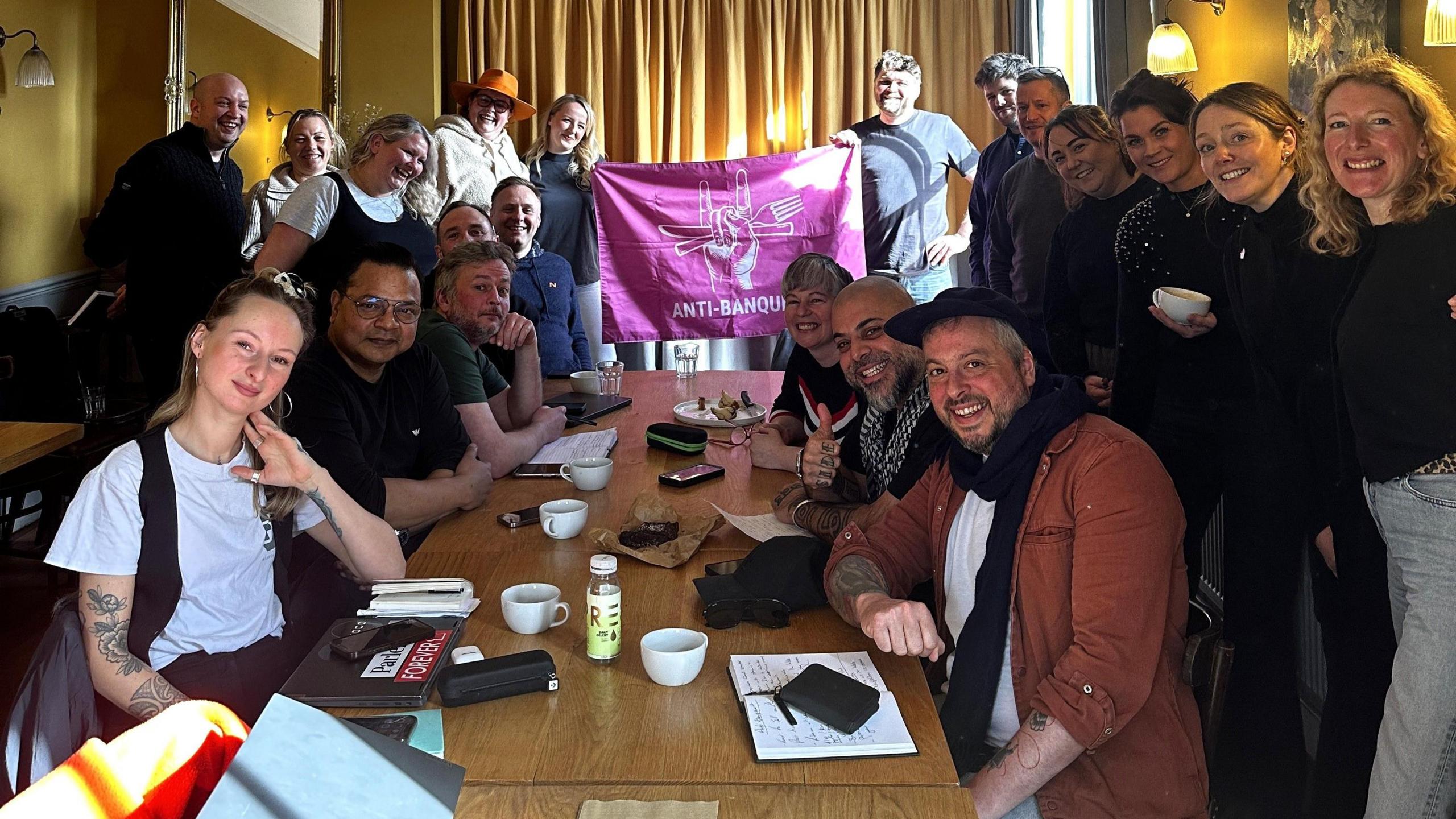 A large group of people sit around a long wooden table and smile at the camera, all of them in casual clothes. They are the organisers of the Anti-Banquet in Bristol and are a mixture of men and women