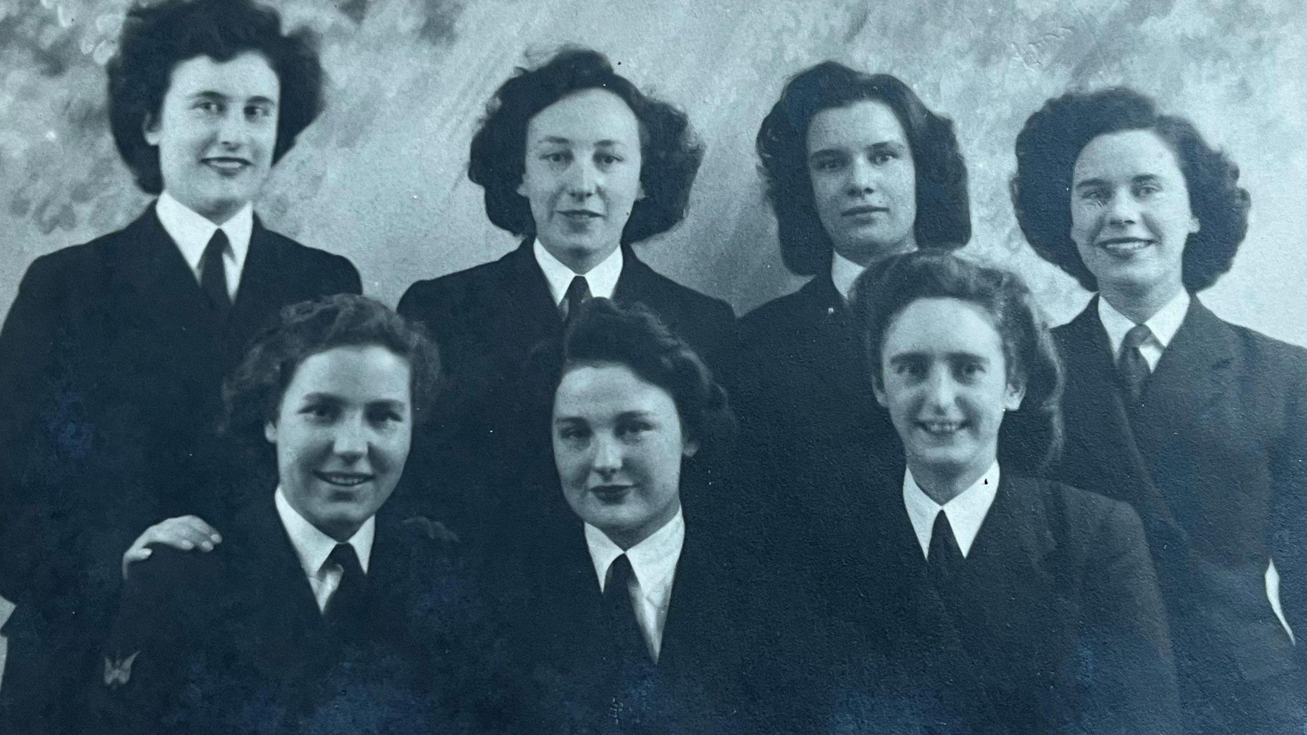 A formal black and white photo of seven women in two rows, four at the back and three at the front. All smiling, with dark hair, smartly dressed in their uniform of white shirt with dark coloured tie and jacket.