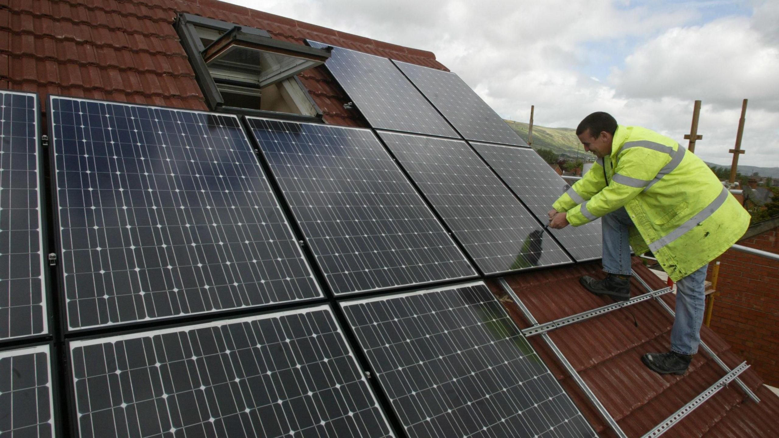 A man wearing a hi-vis jacket fixing dark solar panels to a slate roof.