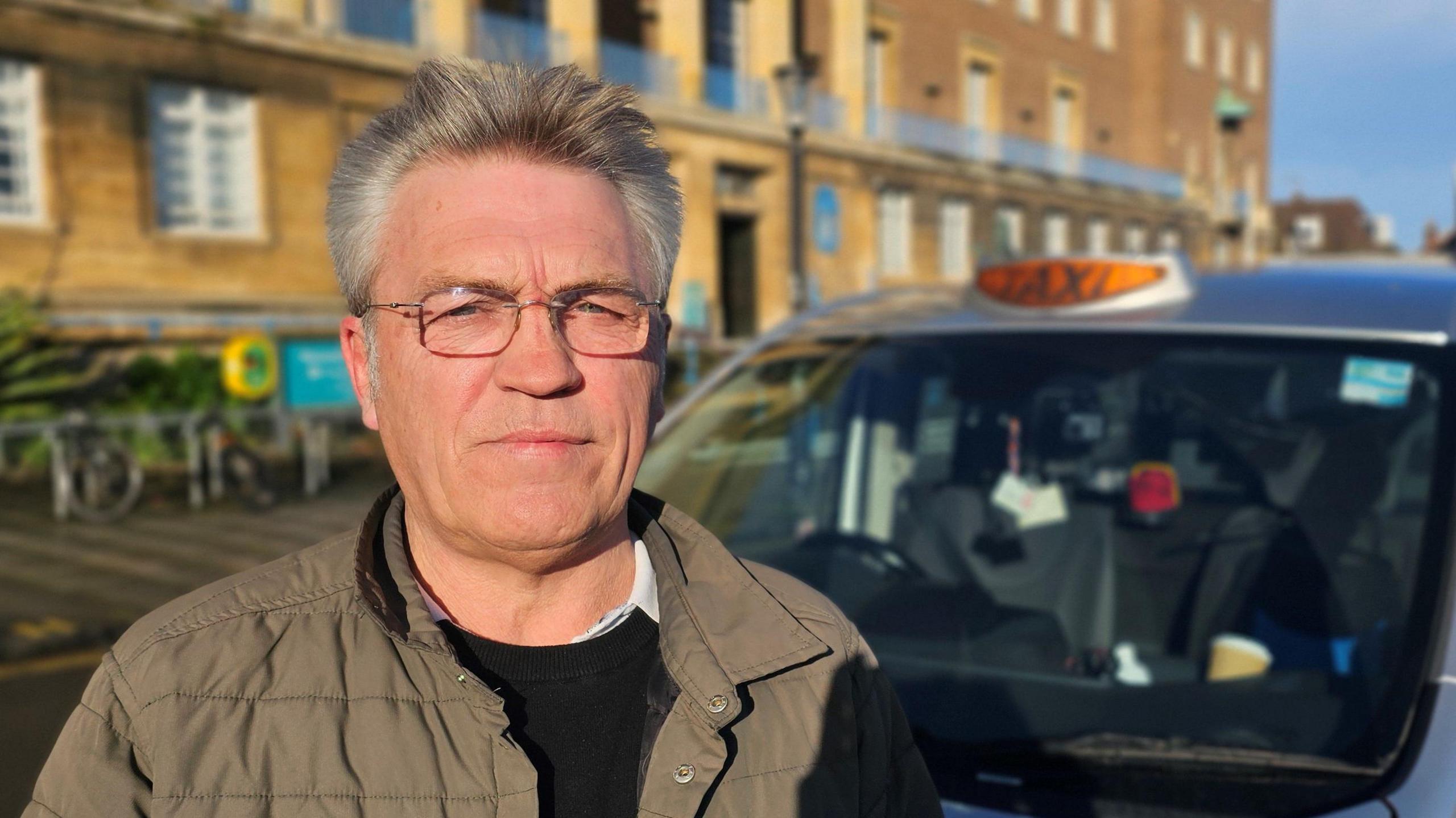 Craig Dimbleby standing outside Norwich City Hall, with his silver cab behind him. He is wearing glasses and a green jacket over a black sweater and white shirt.