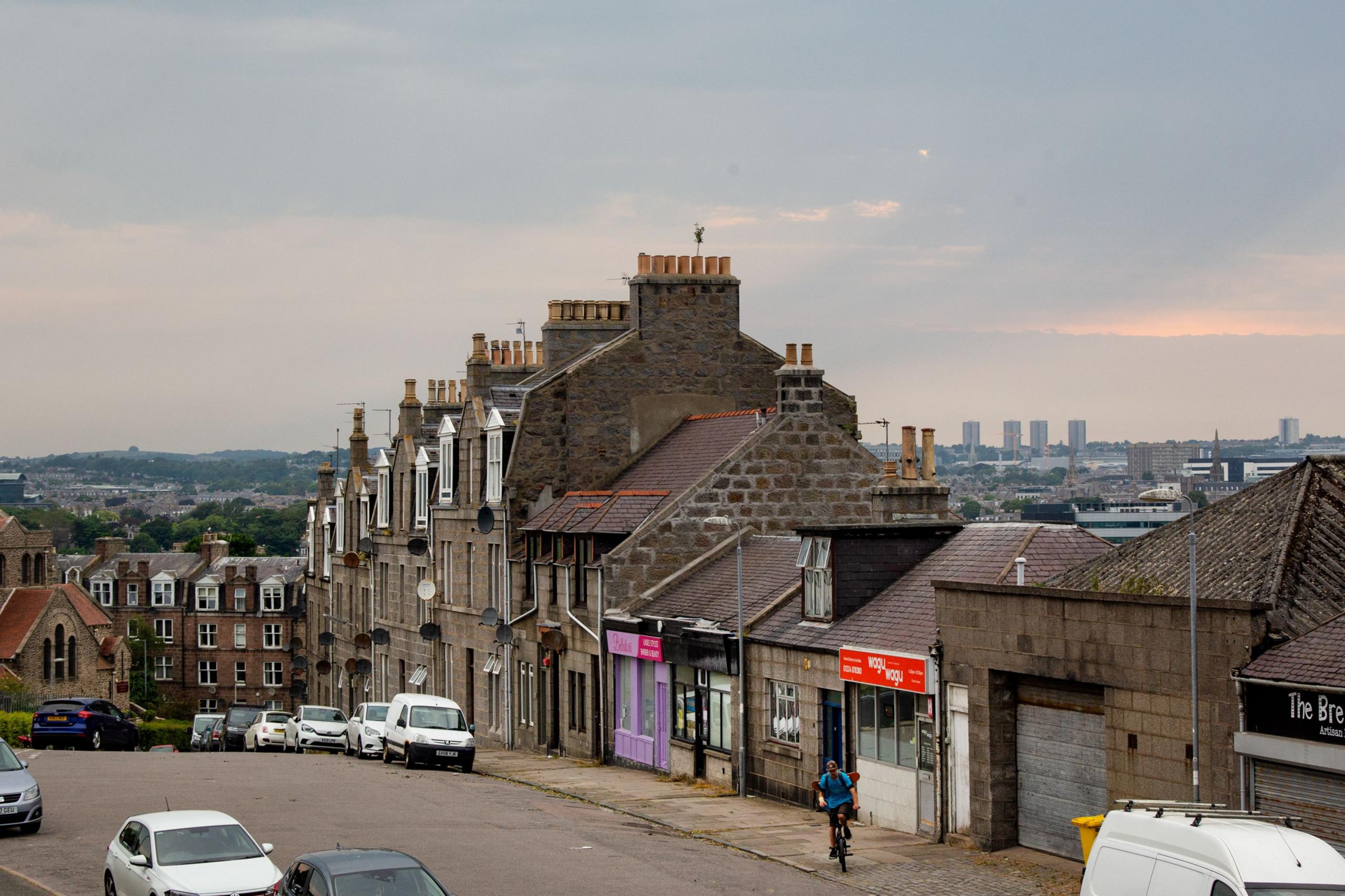 An image of a street in Torry, Aberdeen. We see terraced flats and a row of shops with mixed heights. There are hills and high rise flats in the background.