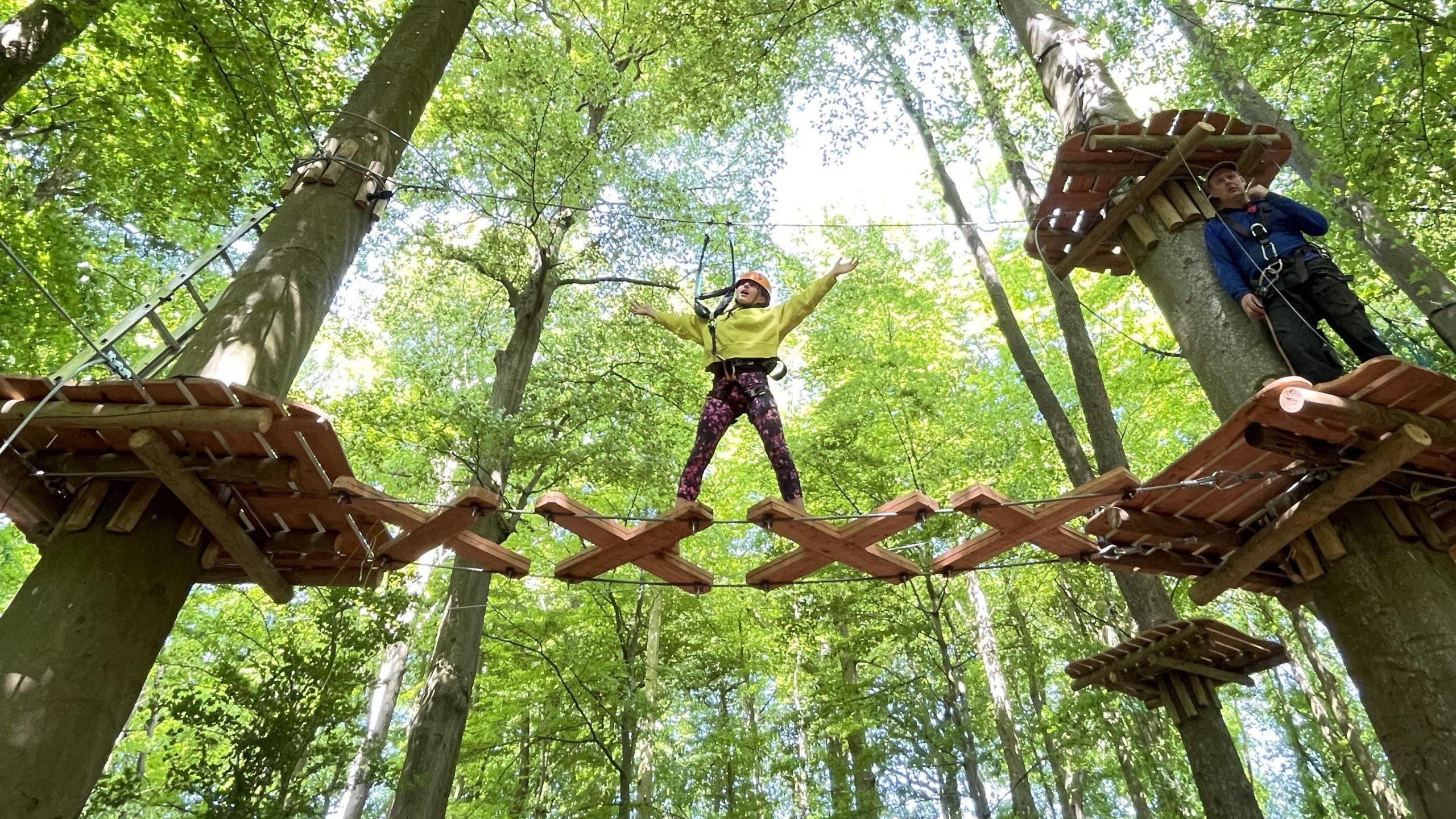 A young girl with black trousers and a yellow top crosses between two trees using a wooden obstacle course as part of an adventure area at Ashton Court in Bristol. In the background are more trees, their leaves showing up light green in the sunshine