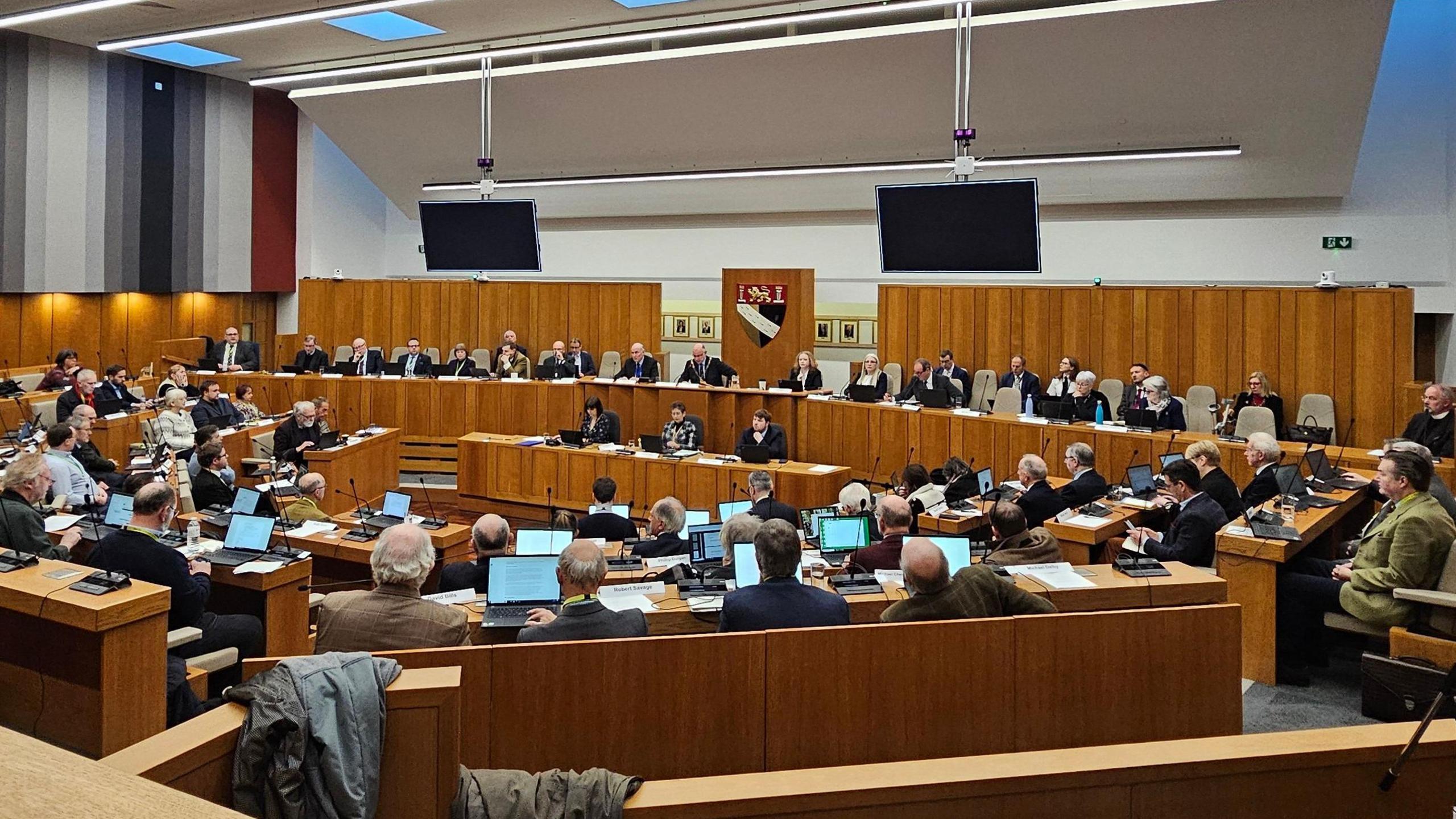 A view of a full council meeting at Norfolk's County Hall. The council chamber is wood panelled, with most seats filled by councillors.