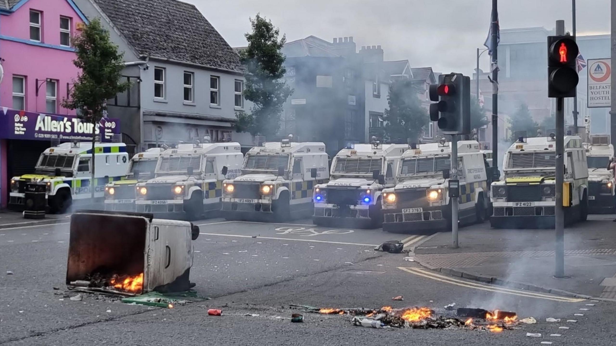 A bin on fire in front of a line of armoured police vehicles in Belfast