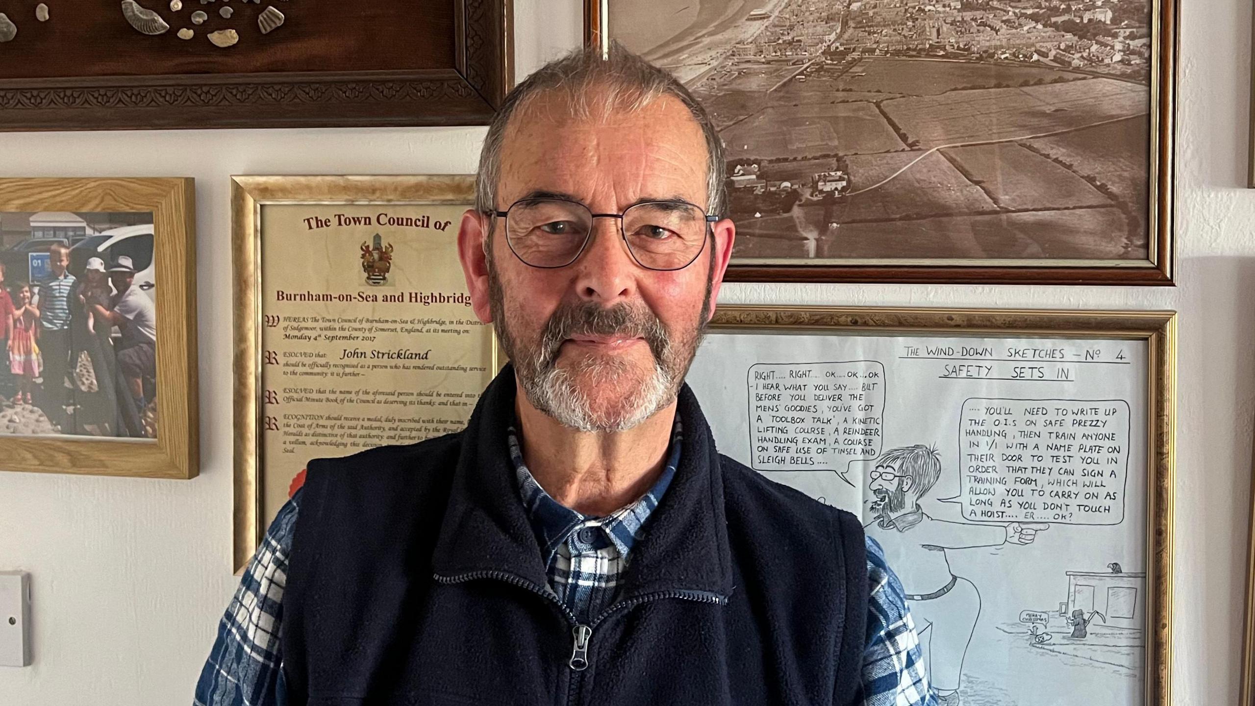 John Strickland is standing in front of a wall with framed photos and documents. He is wearing a checked shirt and a blue vest. 