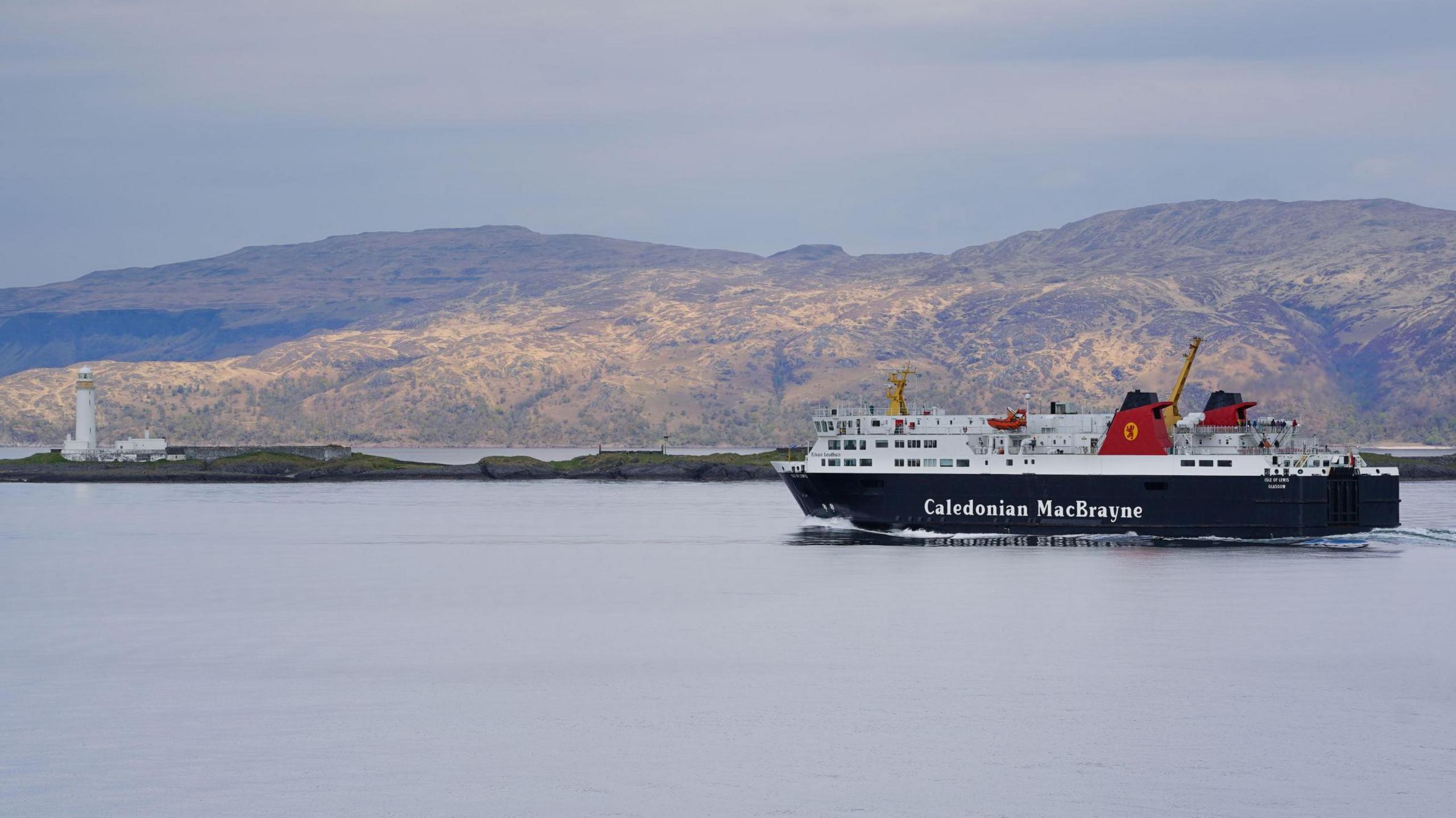 The MV Isle of Lewis is a large black and white ferry with two red funnels either side of each other and yellow and red logos. The ship is sailing in calm waters past low rugged hills.