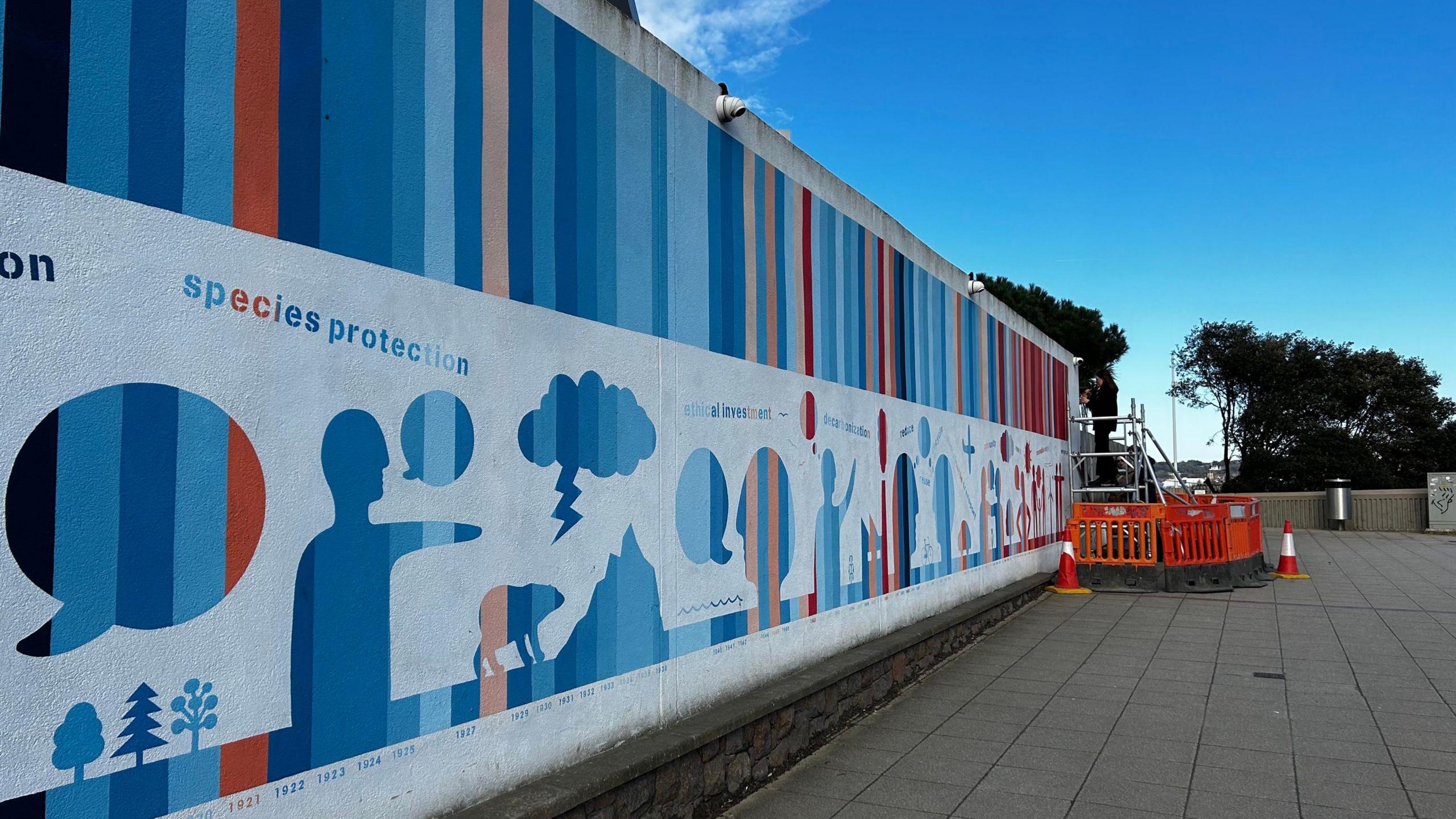 A wall with blue, dark red and orange stripes at the top of it. There is orange barriers at the end of the wall and trees in the distance. 