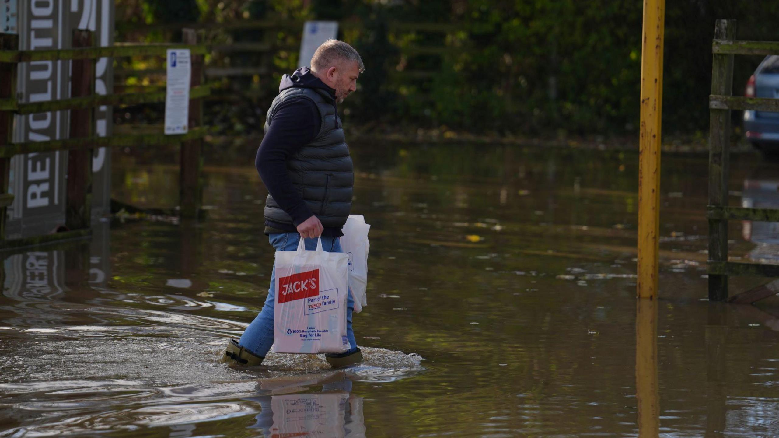 A man walking through knee-high water in a car park carrying two white plastic bags. The bag nearest the camera has "Jack's" written on it and we can see small bow waves from the front of his wellies as he moved.