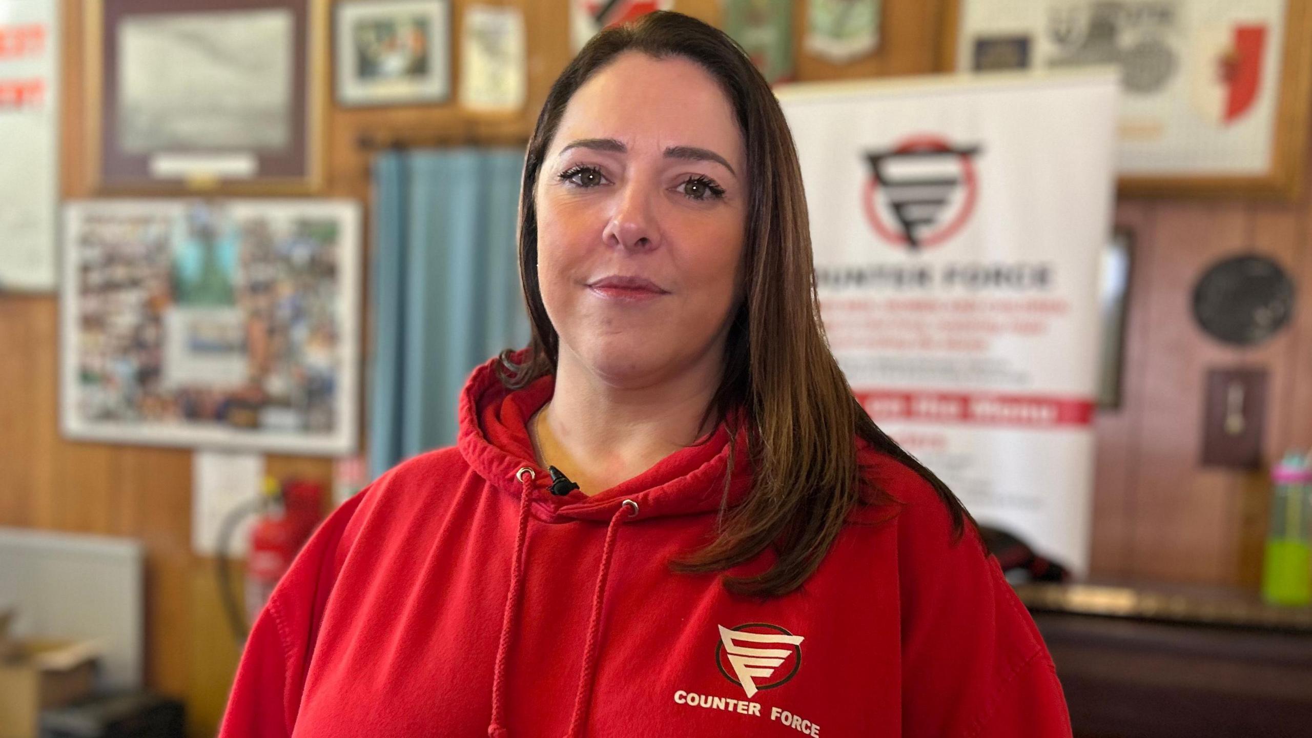 Emma Port yn gwisgo siwmper coch gyda logo Counter Force, gwallt hir brown
