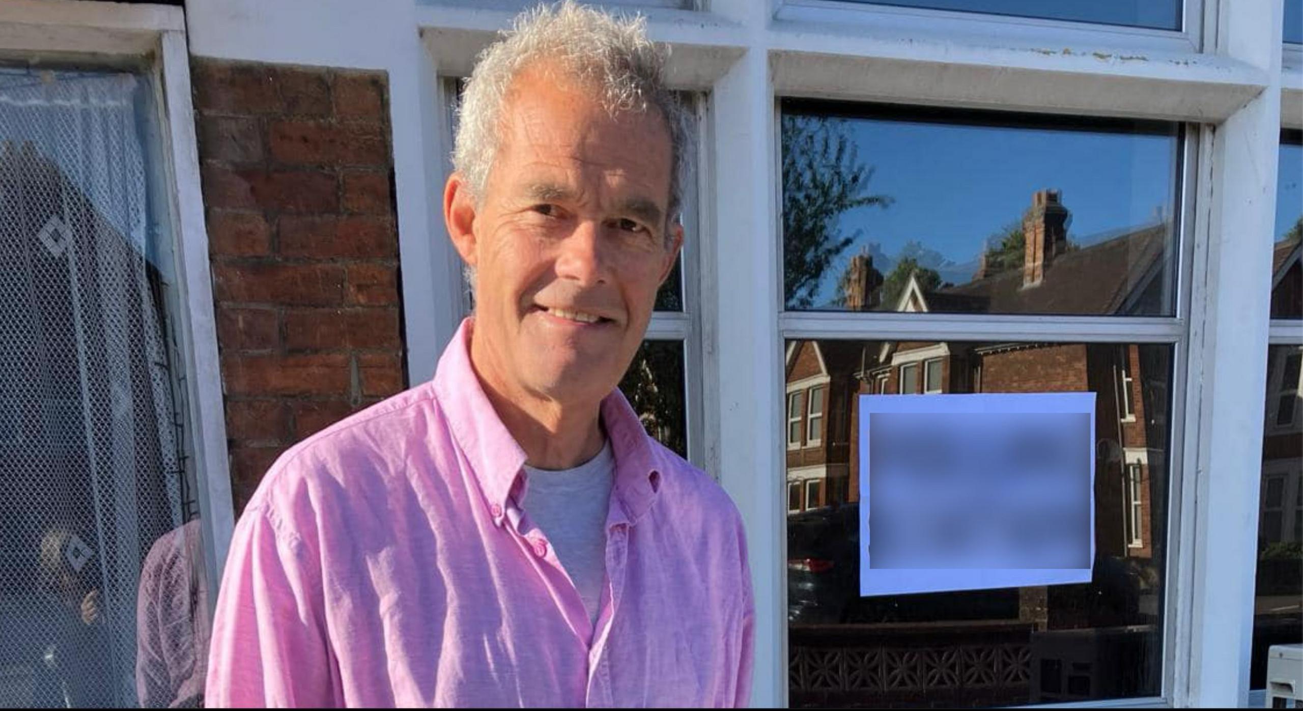 Michael Barber with short white hair smiling at the camera and wearing a pink shirt over a white T-shirt. He is standing in front of white framed windows, one of which has a white notice pinned to it.