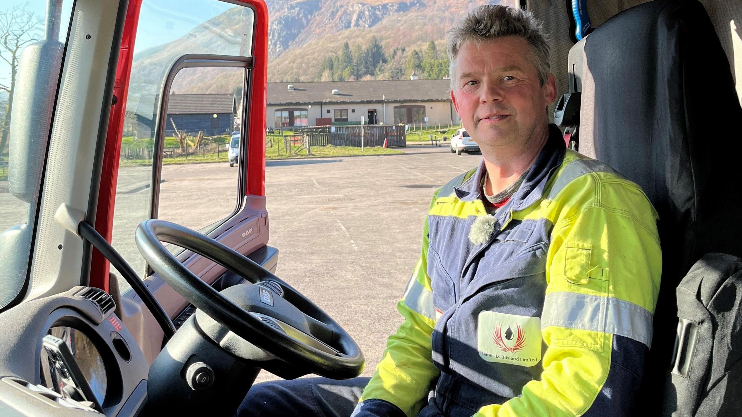 Stuart Youens sitting in a lorry cab wearing a grey and yellow boiler suit. Through the open door there is a car par, a house and some hills in the background.