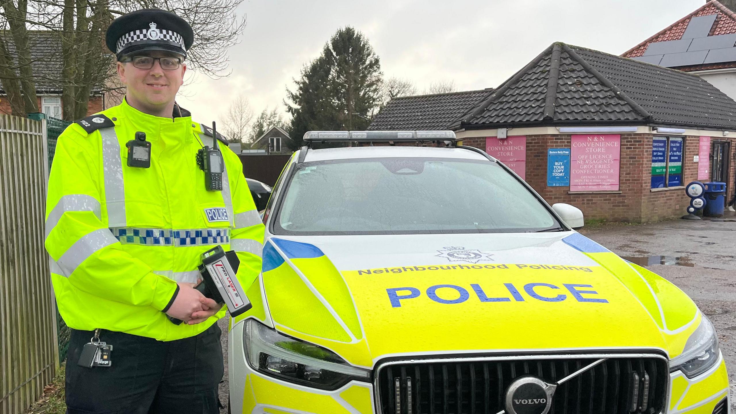 PC Daniel Brock stands dressed in high vis and police hat. He is holding a speed gun and is standing next to his marked police car. 