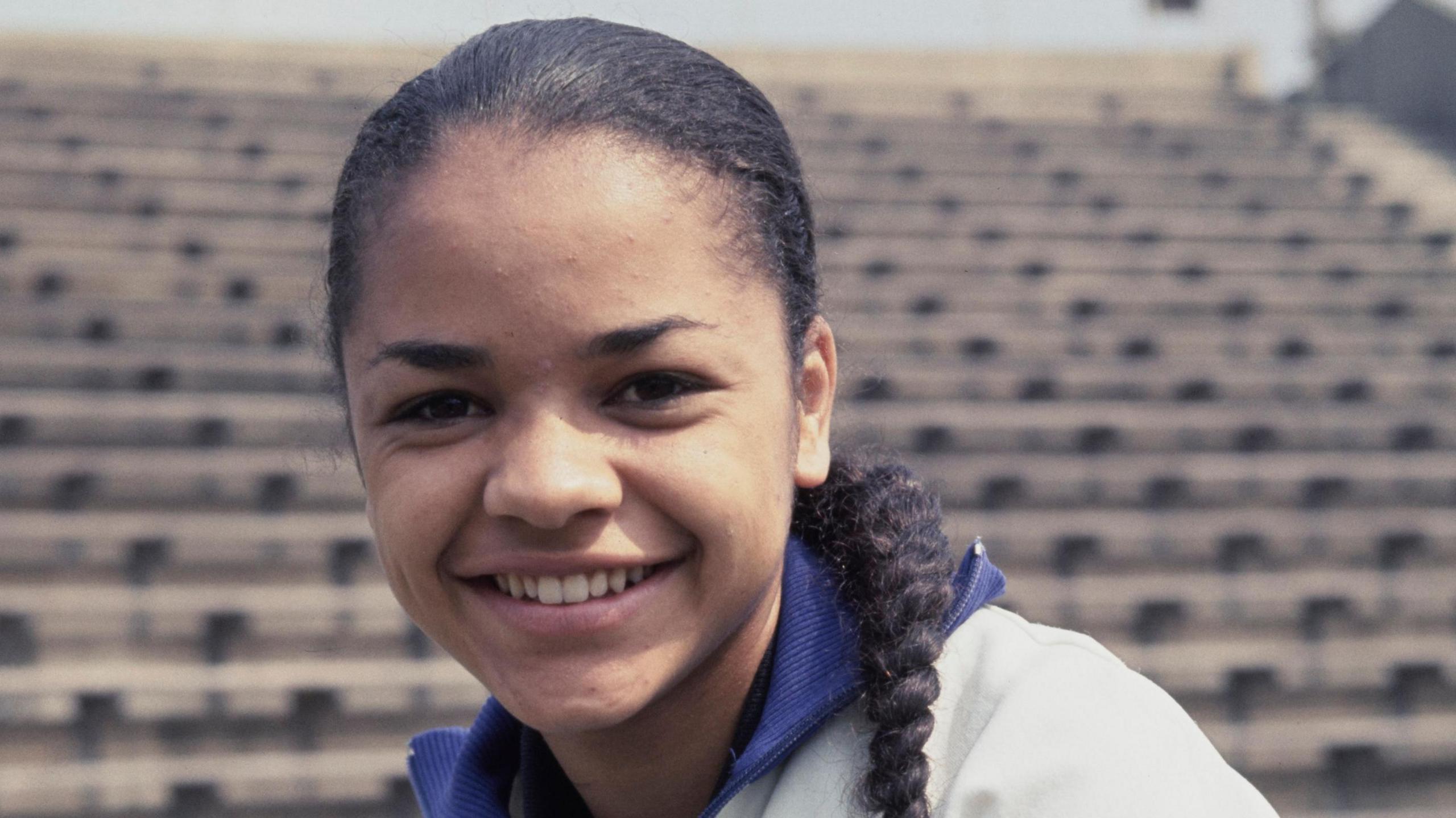 A portrait of sprinter Anita Neil taken in June 1967 at the Crystal Palace National Sports Centre. Ms Neil, who is wearing a sports top and has her hair in a plait, is smiling.