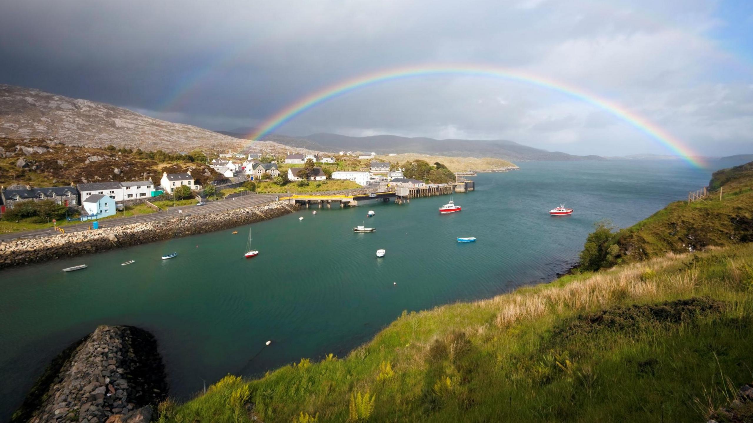 A Hebridean scene with houses on a rocky shoreline and a rainbow arching over a bay with boats dotted across the surface of the sea.