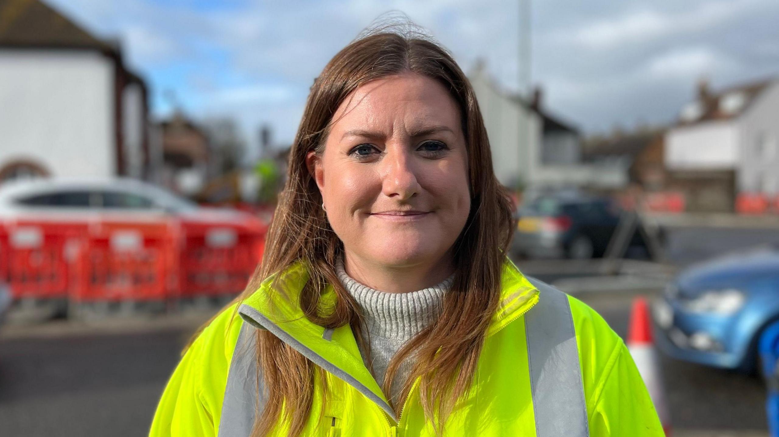 Andrea smiling at the camera wearing a high-vis with traffic behind her. She has medium length brown hair and a turtle neck jumper