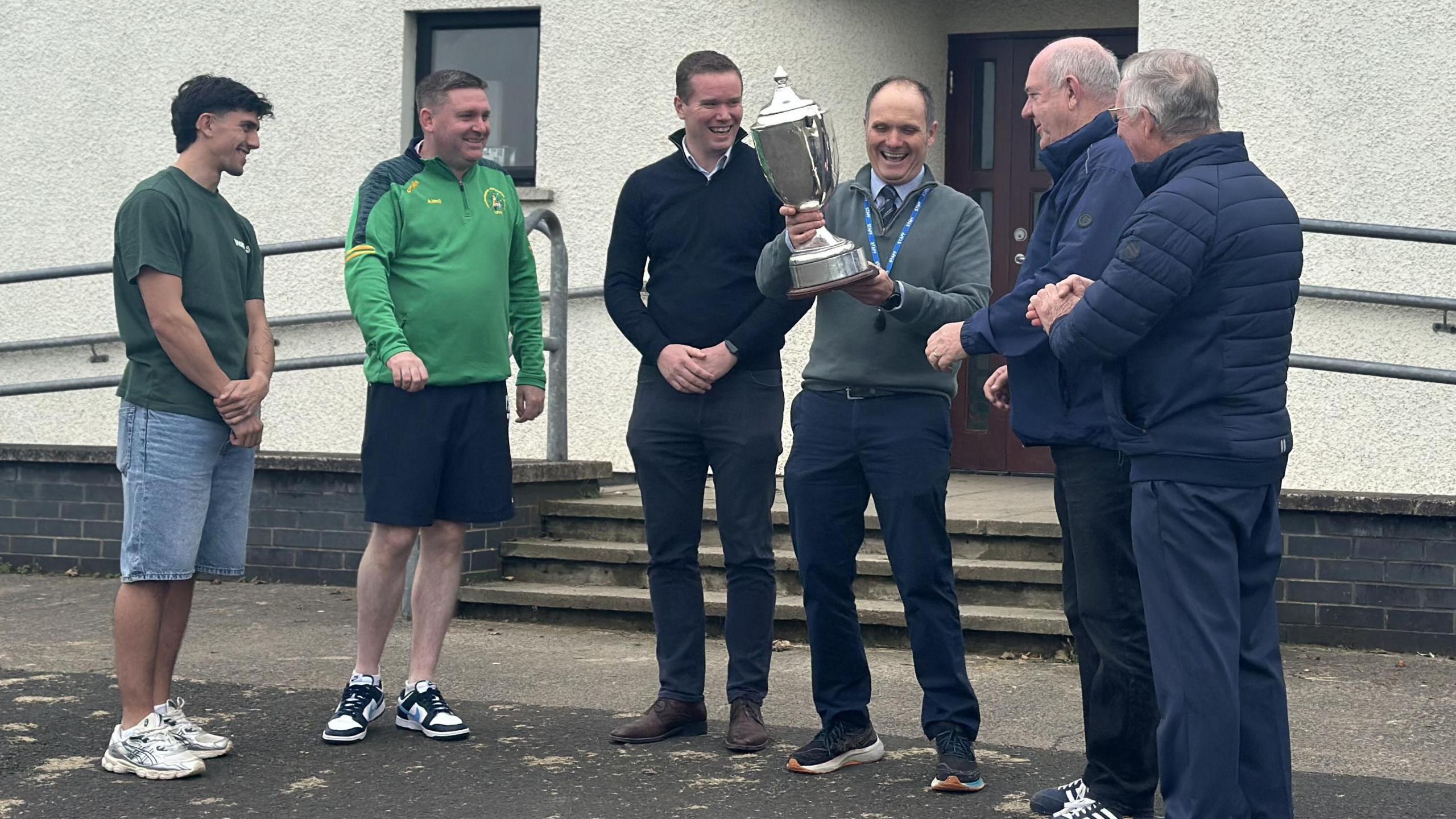 Six men stand outside a grey building. They are all smiling and one man is holding a silver trophy.