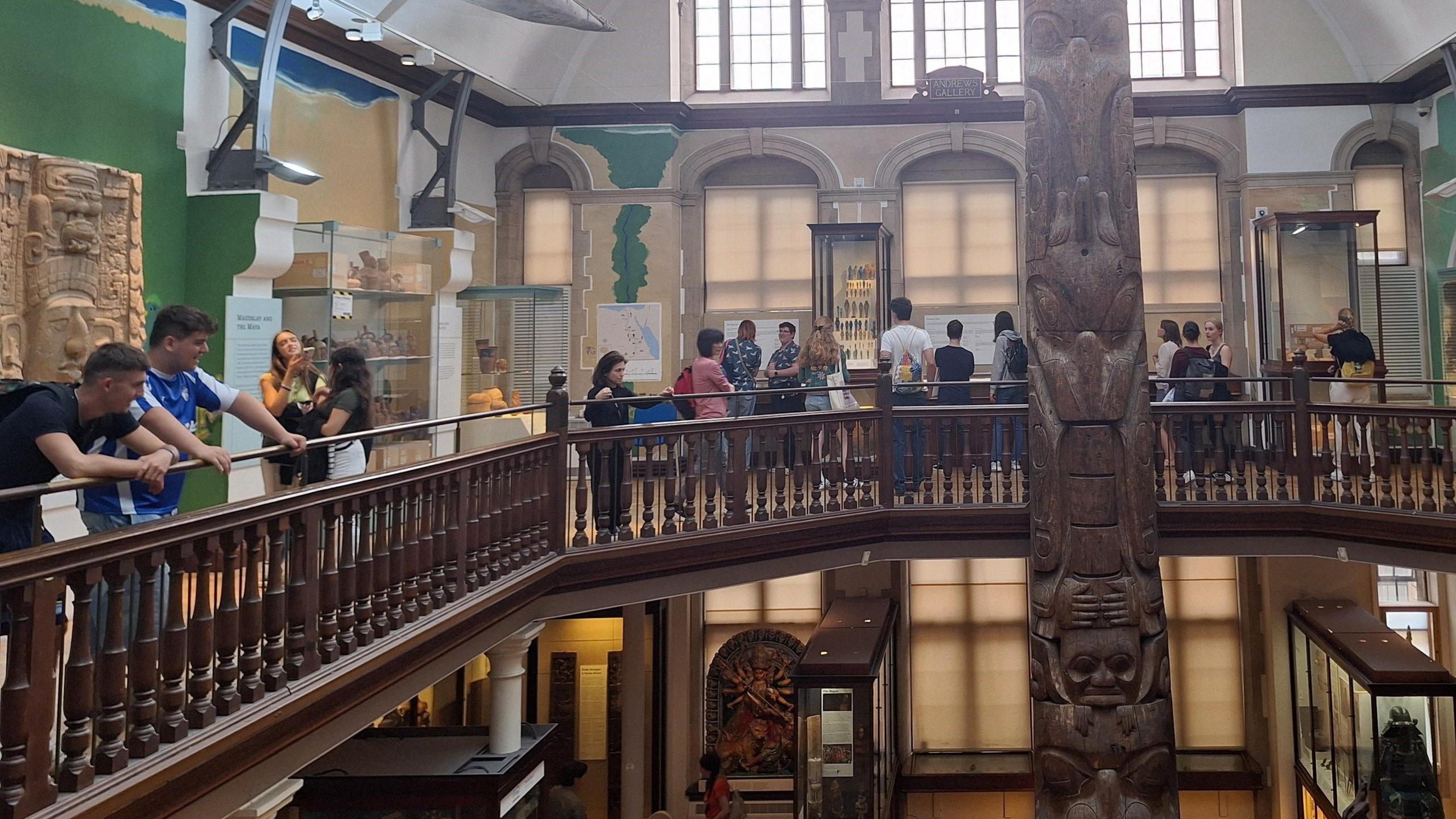 A number of people in the Museum of Archaeology and Anthropology, which shows a wooden mezzanine floor, with displays and a wooden pole in the middle.