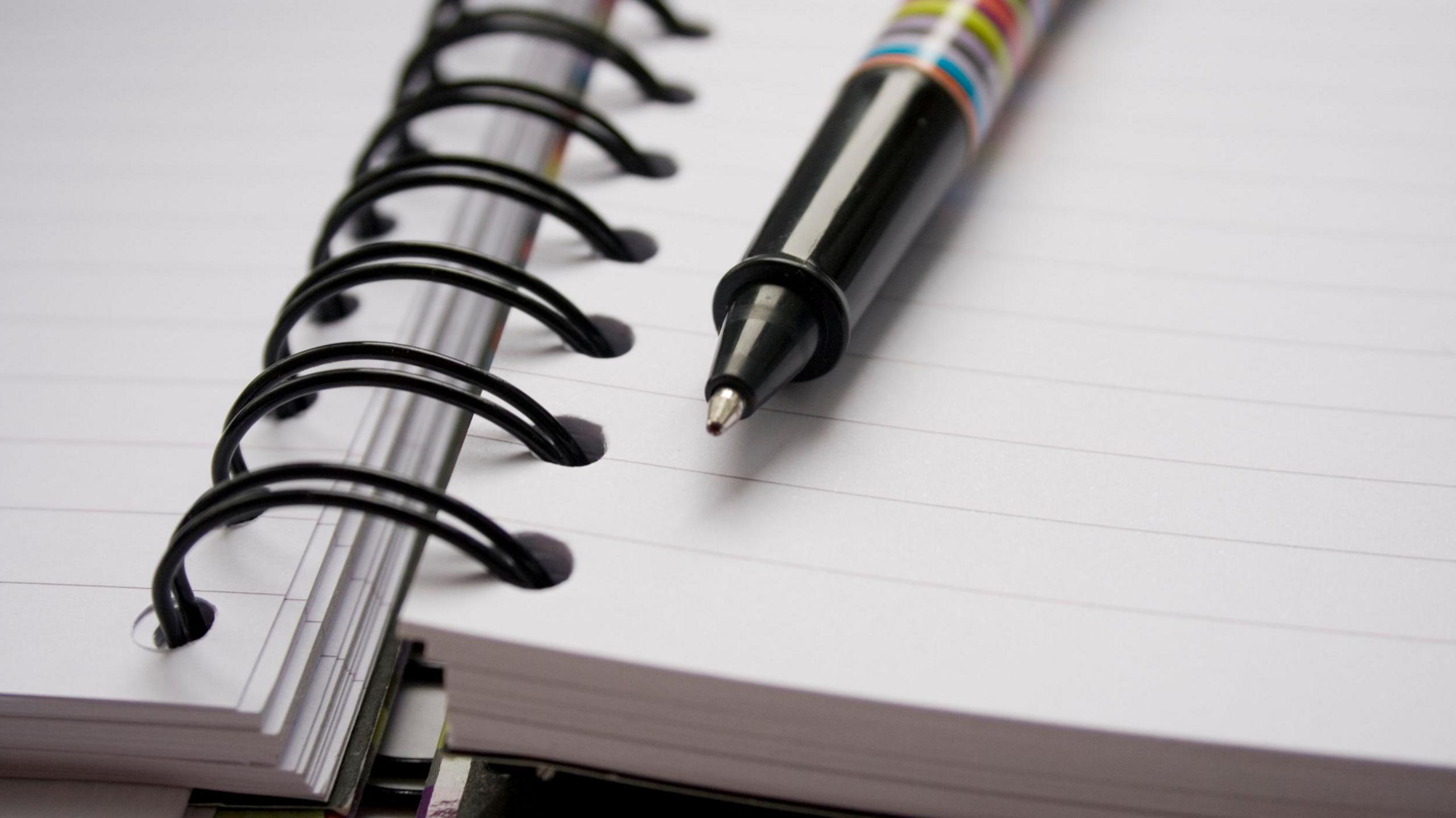 Close-up of an open spiral-bound notebook with lined pages. A diagonally placed pen featuring a colourful striped design rests on the notebook.