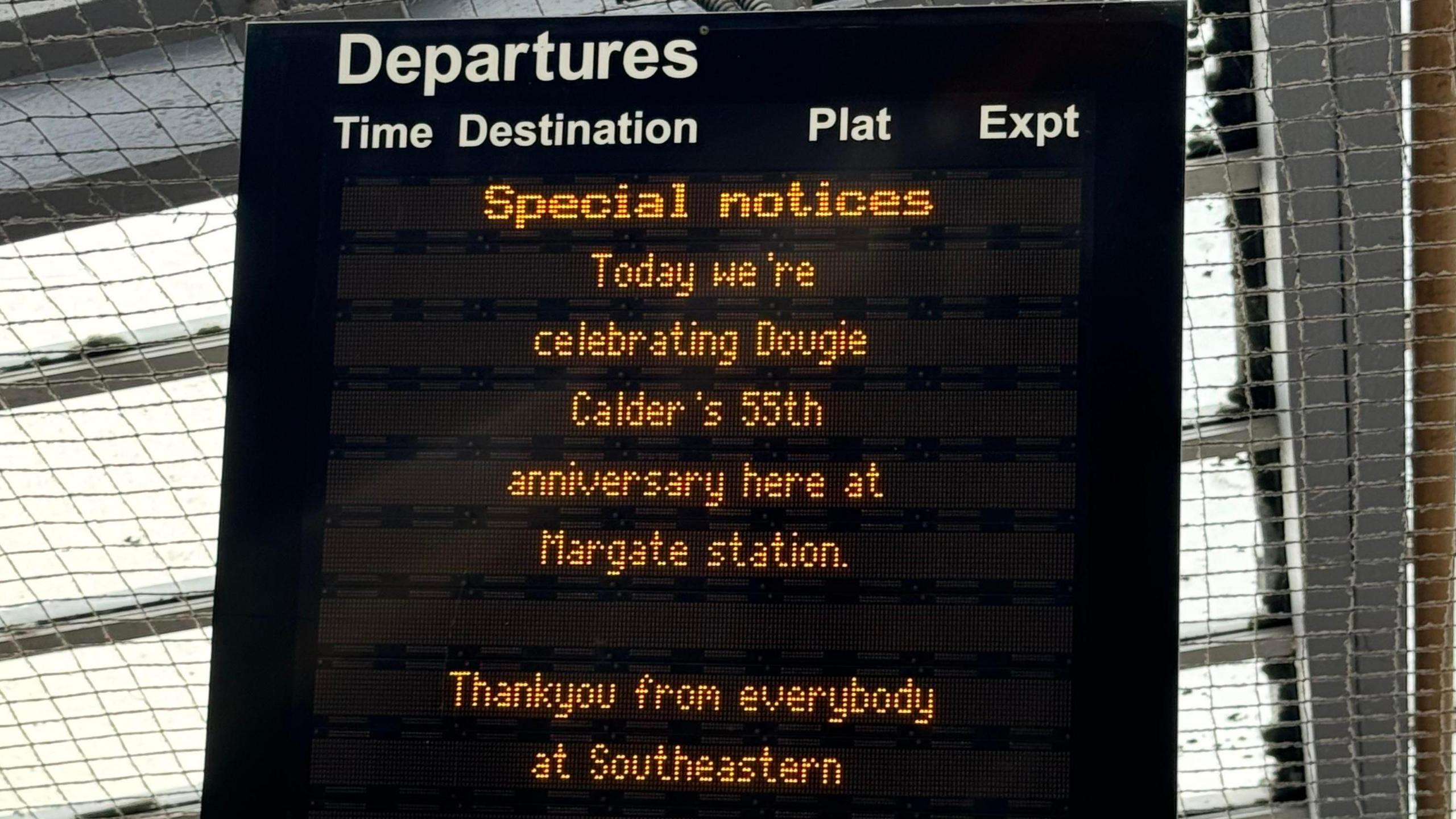 A black digital train notice board. In orange writing it reads 'Special notices. Today we're celebrating Dougie Calder's 55th anniversary here at Margate Station. Thankyou from everybody at Southeastern.' 