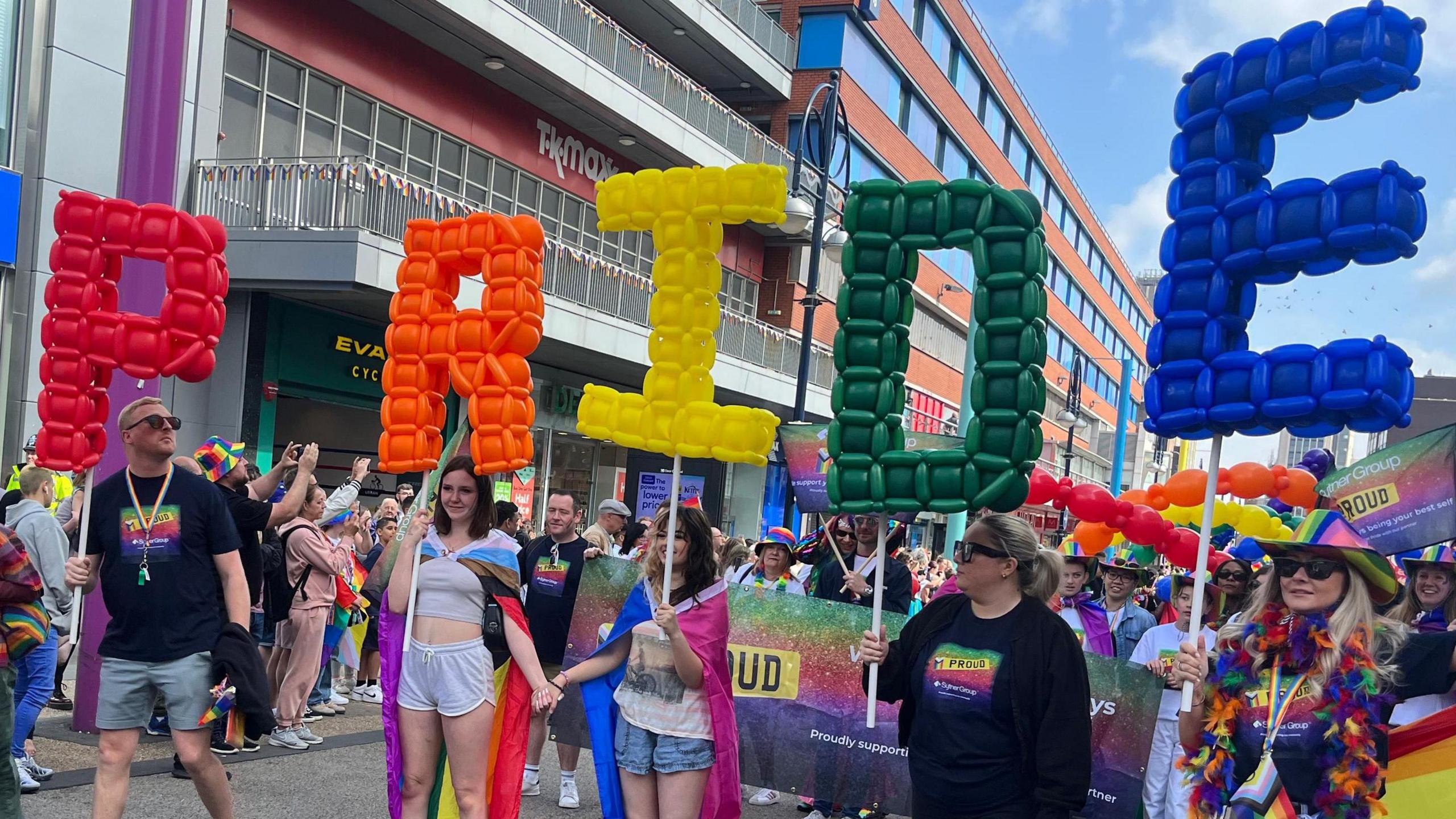 People holding up inflatable letters spelling out Pride with a large crowd behind them.