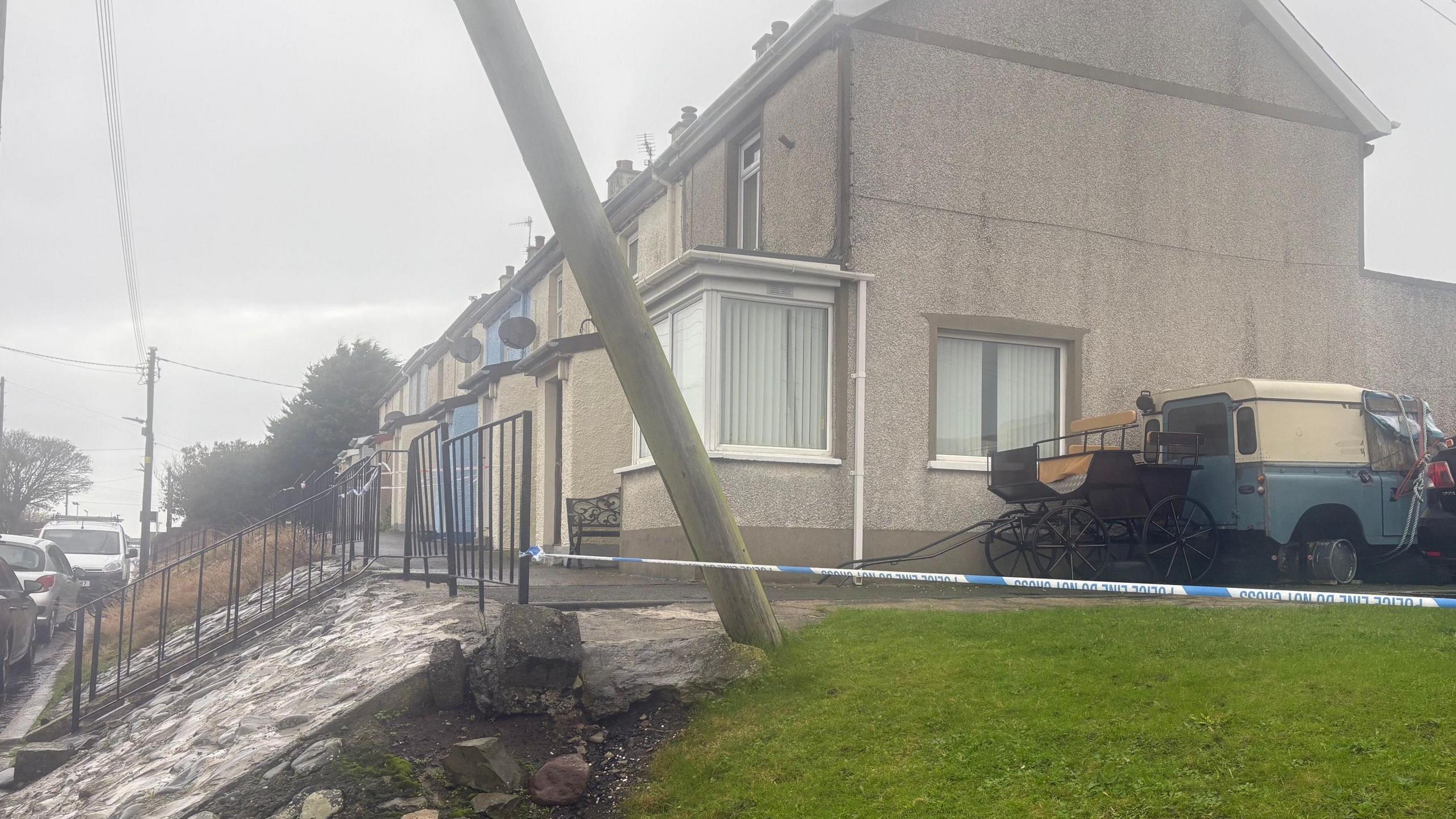 A row or terraced houses with steps leading up to them and cars parked in front. A police cordon is taped to the railing.
