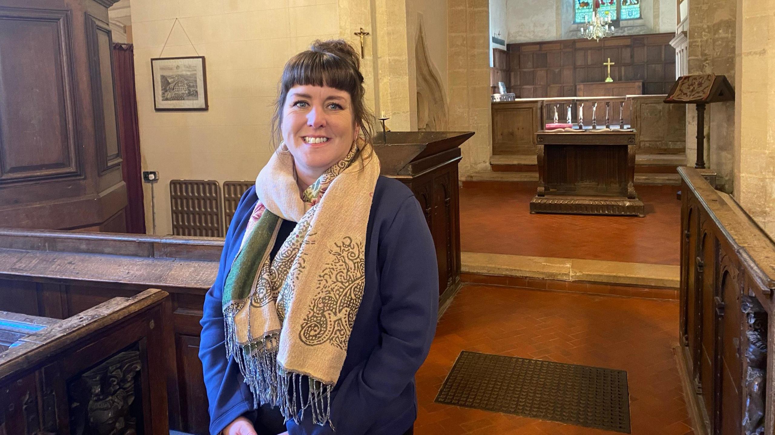 Marie Sellers, pictures inside St Kenelm's. She has dark hair pulled back into a bun, and a short fringe, and wears a blue cardigan and a large, light-coloured scarf wrapped around her neck. She is smiling at the camera next to one of the pews. 