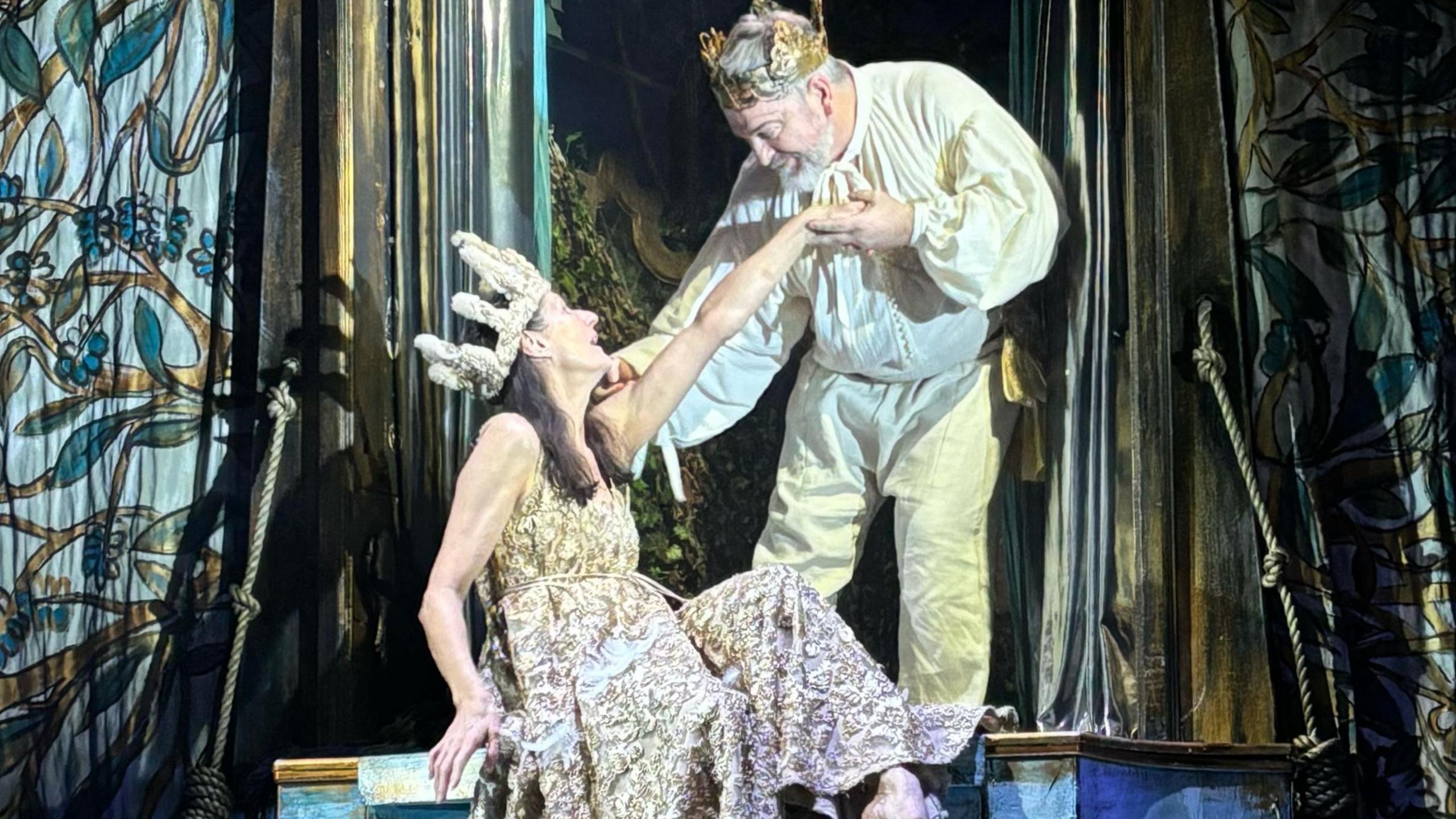 Two dancers wearing light-coloured clothing with the woman sitting down with her hand held by a man who is standing up and leaning down towards her and both are wearing crowns